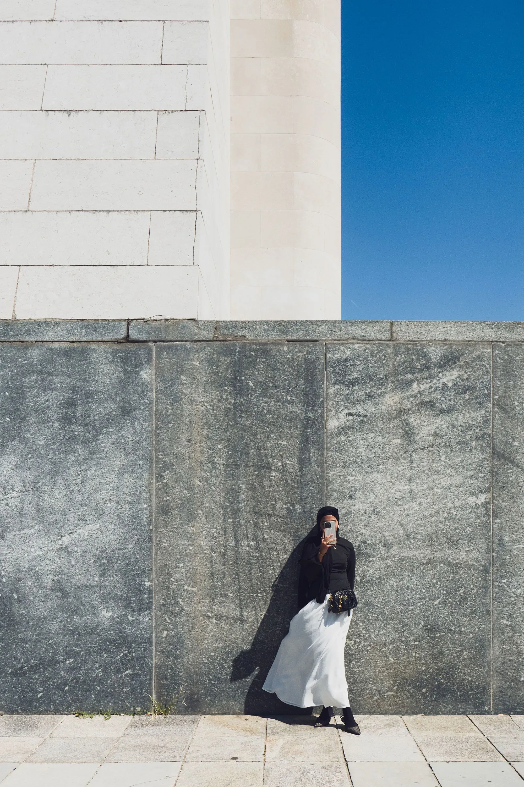 امرأة ترتدي فستانًا أبيض وسروالًا أسود، تتكئ على جدار رمادي، تحمل كاميرا في يدها، تحت سماء زرقاء.