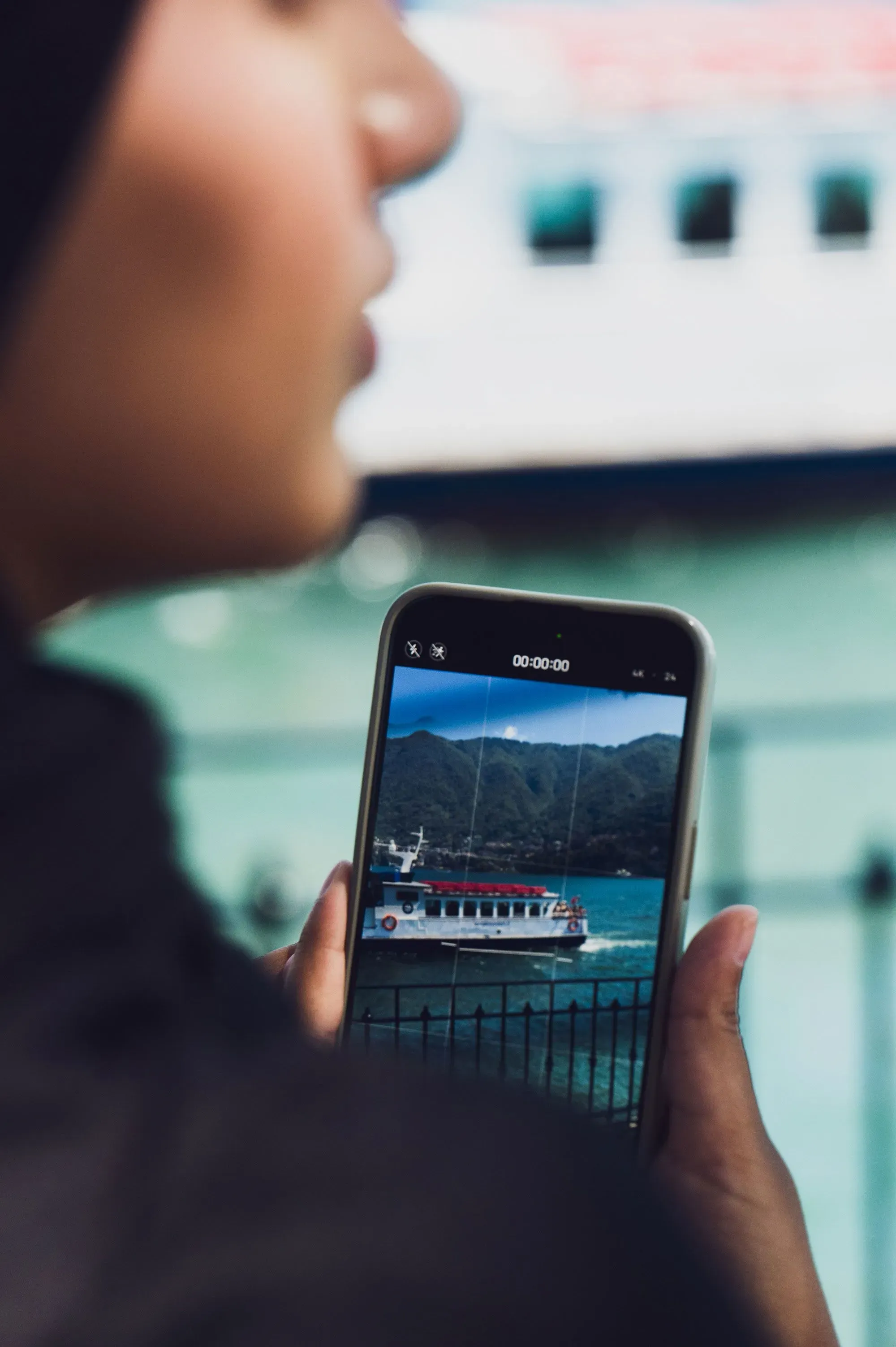 امرأة ترتدي حجابًا تحمل هاتفًا محمولًا، تلتقط صورة لقارب يبحر في المياه مع جبال في الخلفية، مما يعكس جمال الطبيعة.