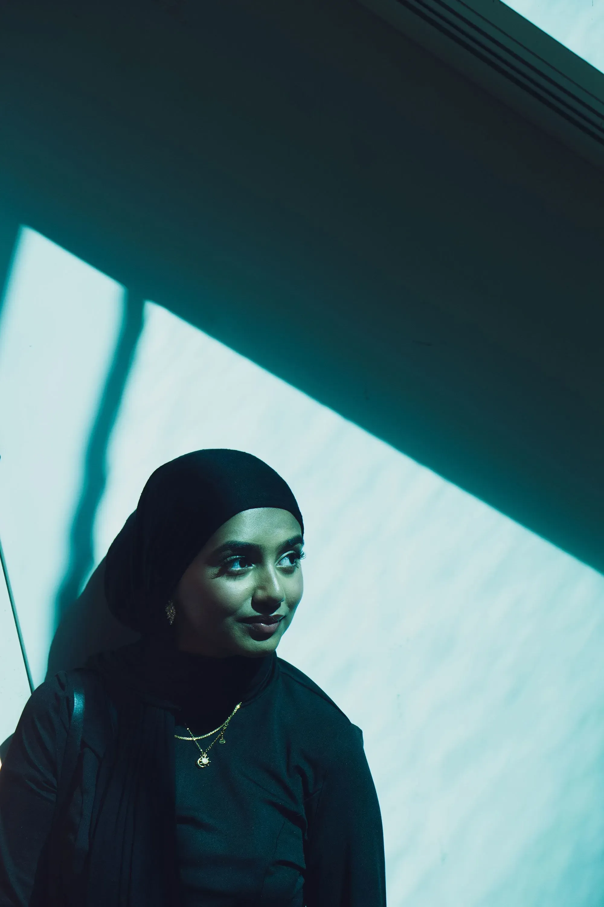 امرأة ترتدي حجابًا أسود وتبتسم بينما تتلقى الضوء من نافذة، مما يخلق ظلالًا جميلة، تعكس نشاطها في تنظيم حفلات الزفاف.