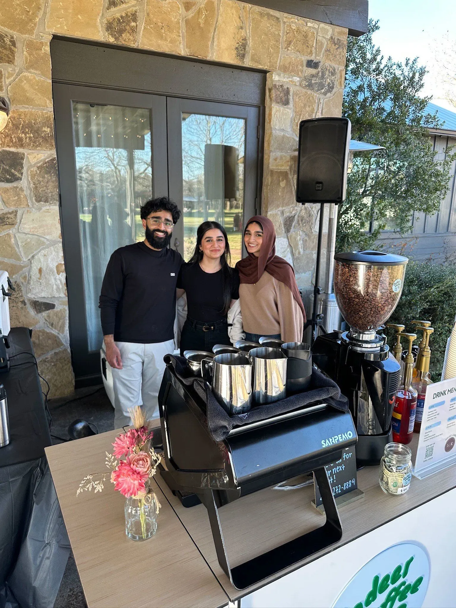 ثلاثة أشخاص يقفون أمام كشك تقديم المشروبات في حفل زفاف، مع معدات تقديم، وزهور، وإضاءة دافئة في الخلفية.