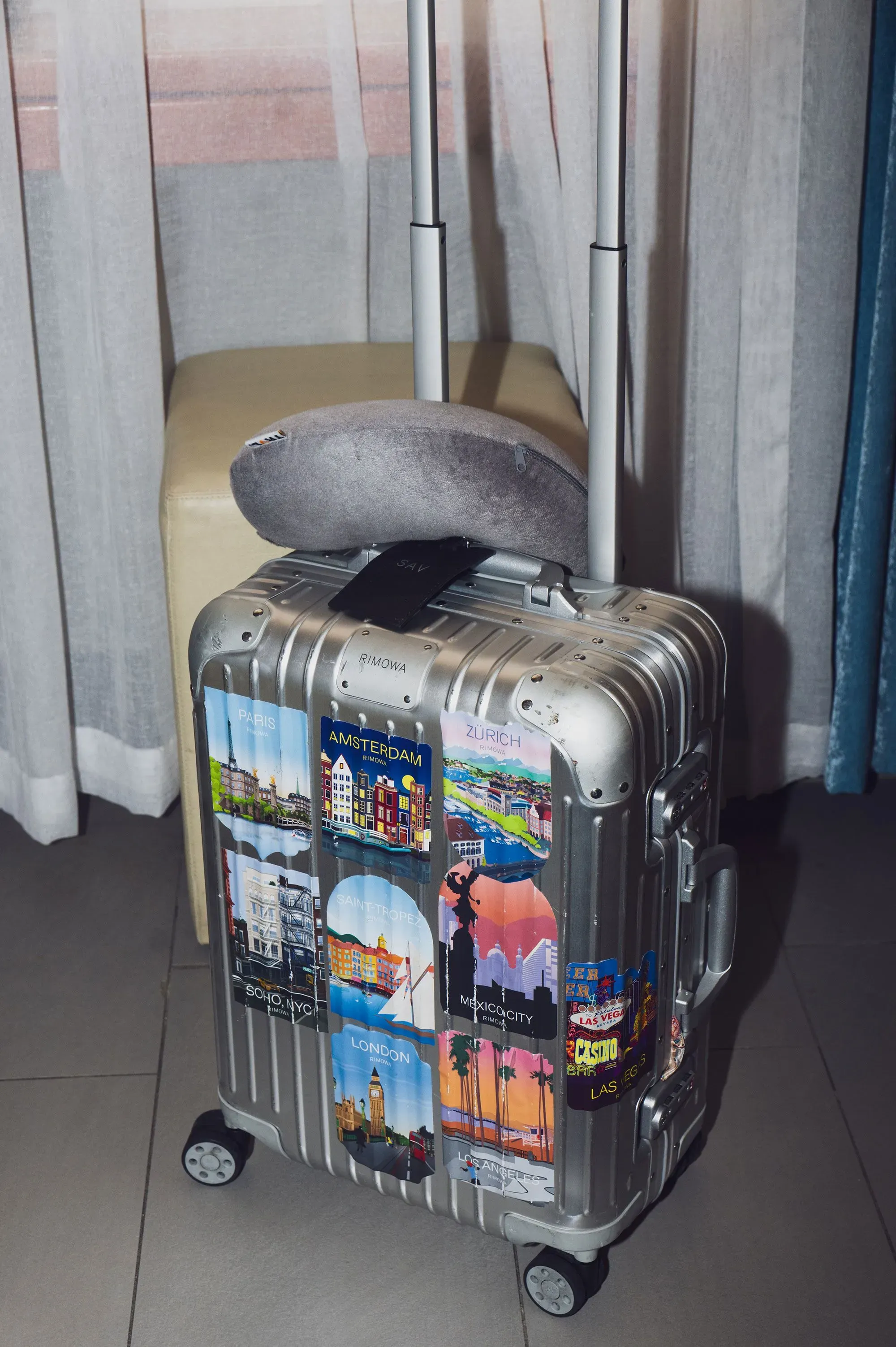 حقيبة سفر فضية تحمل ملصقات لمدن عالمية، موضوعة بجانب كرسي في غرفة داخلية، تعكس أسلوب حياة السفر والمغامرة.