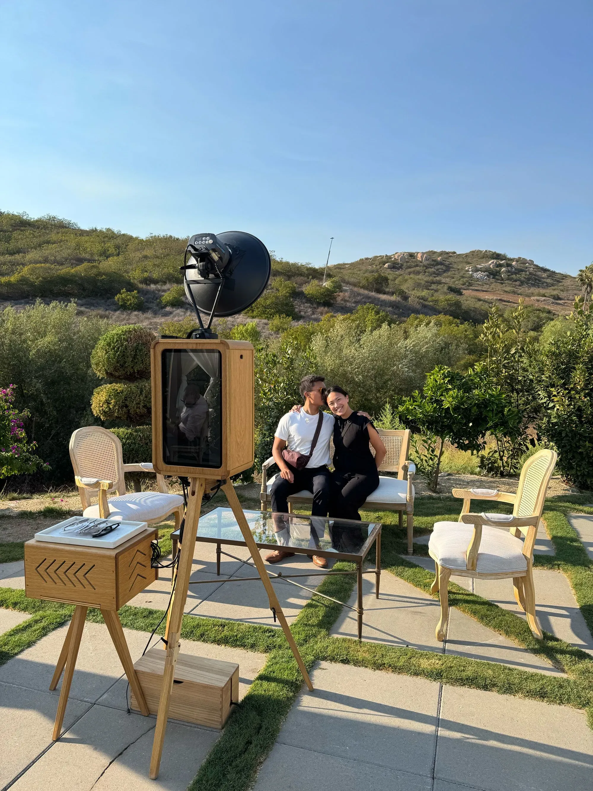 تظهر الصورة شار أغيلار وزوجها يجلسان معًا أمام كشك تصوير خشبي في حفل زفاف، محاطين بمناظر طبيعية جبلية في جنوب كاليفورنيا.