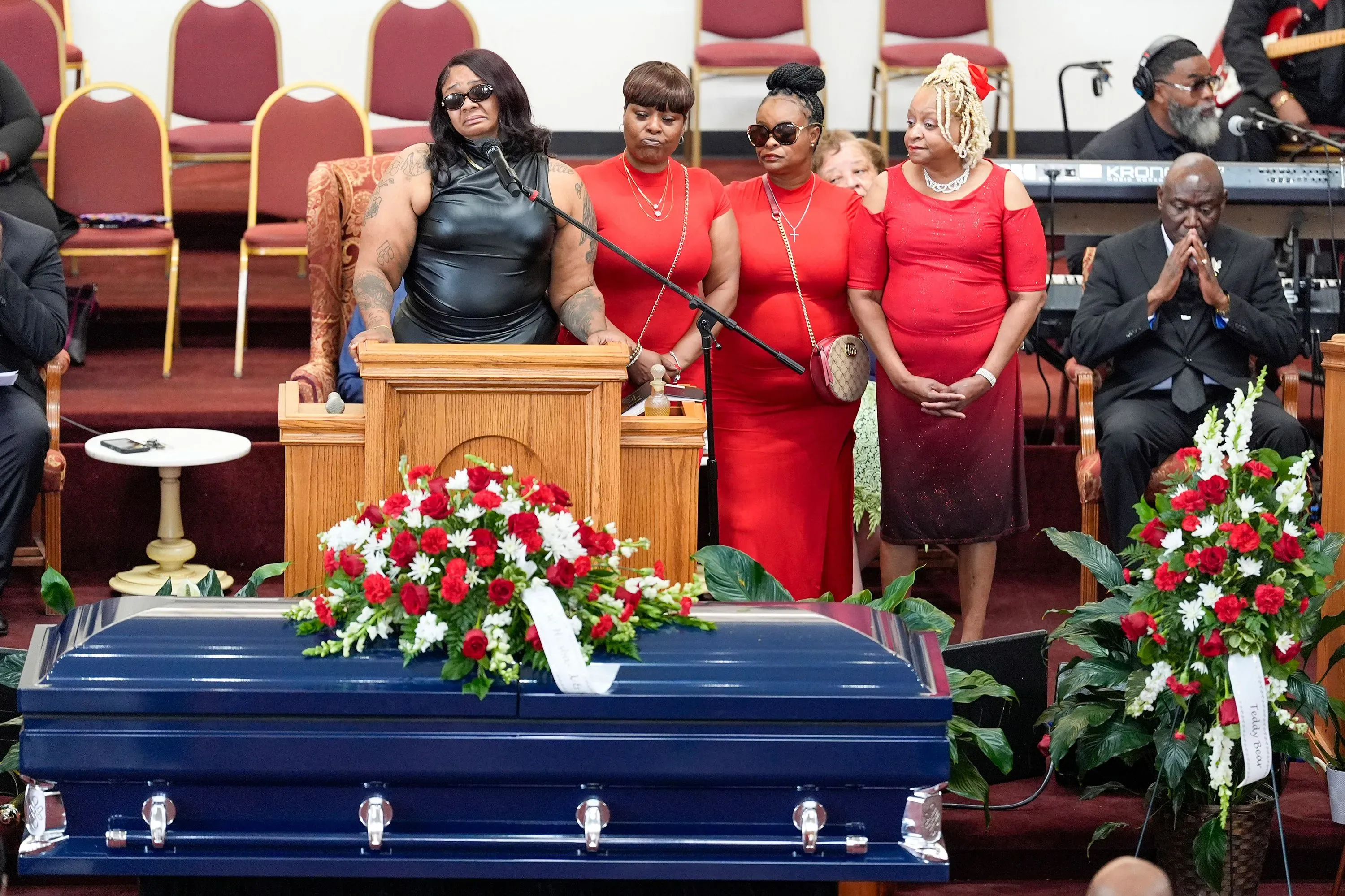 خلال مراسم تأبين، تتحدث امرأة ترتدي سترة جلدية أمام تابوت أزرق مزين بالزهور، بينما تقف أربع نساء يرتدين الأحمر خلفها.