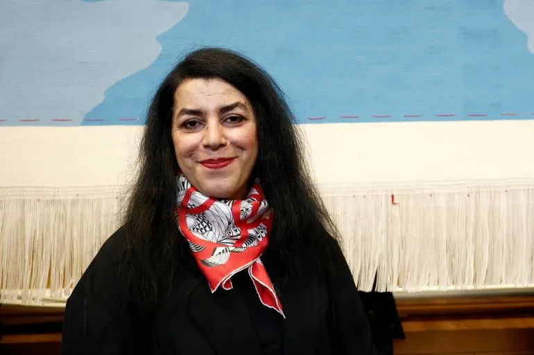 تظهر الكاتبة والمخرجة Marjane Satrapi مبتسمة، ترتدي وشاحاً مزخرفاً، في خلفية فنية تعكس تأثيرها الثقافي وأعمالها الأدبية.