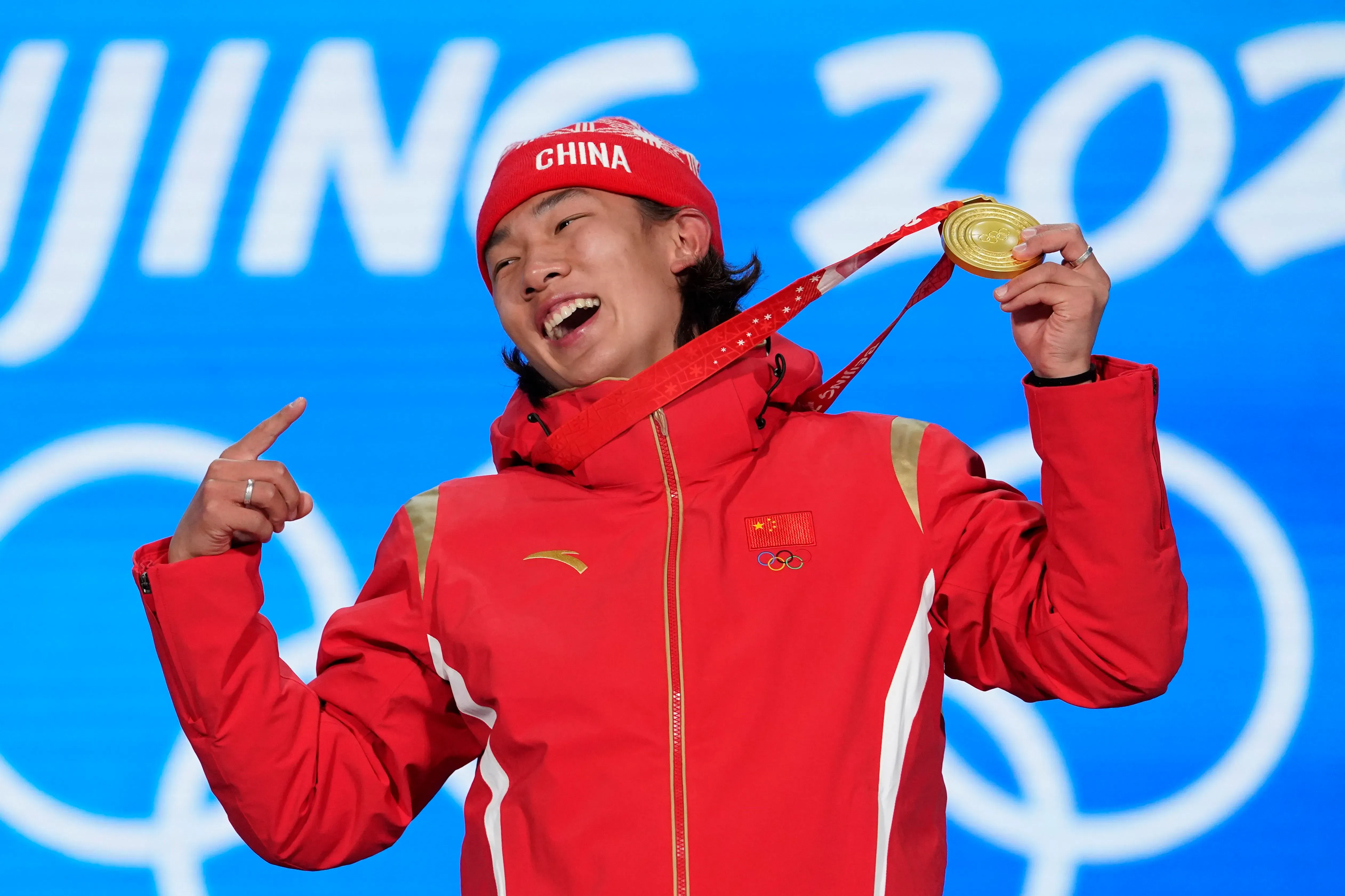 سوي ييمينغ، المتزلج الصيني، يحتفل بفوزه بالميدالية الذهبية في الألعاب الأولمبية الشتوية 2022، مرتديًا زيًا رياضيًا أحمر، مع خلفية زرقاء.