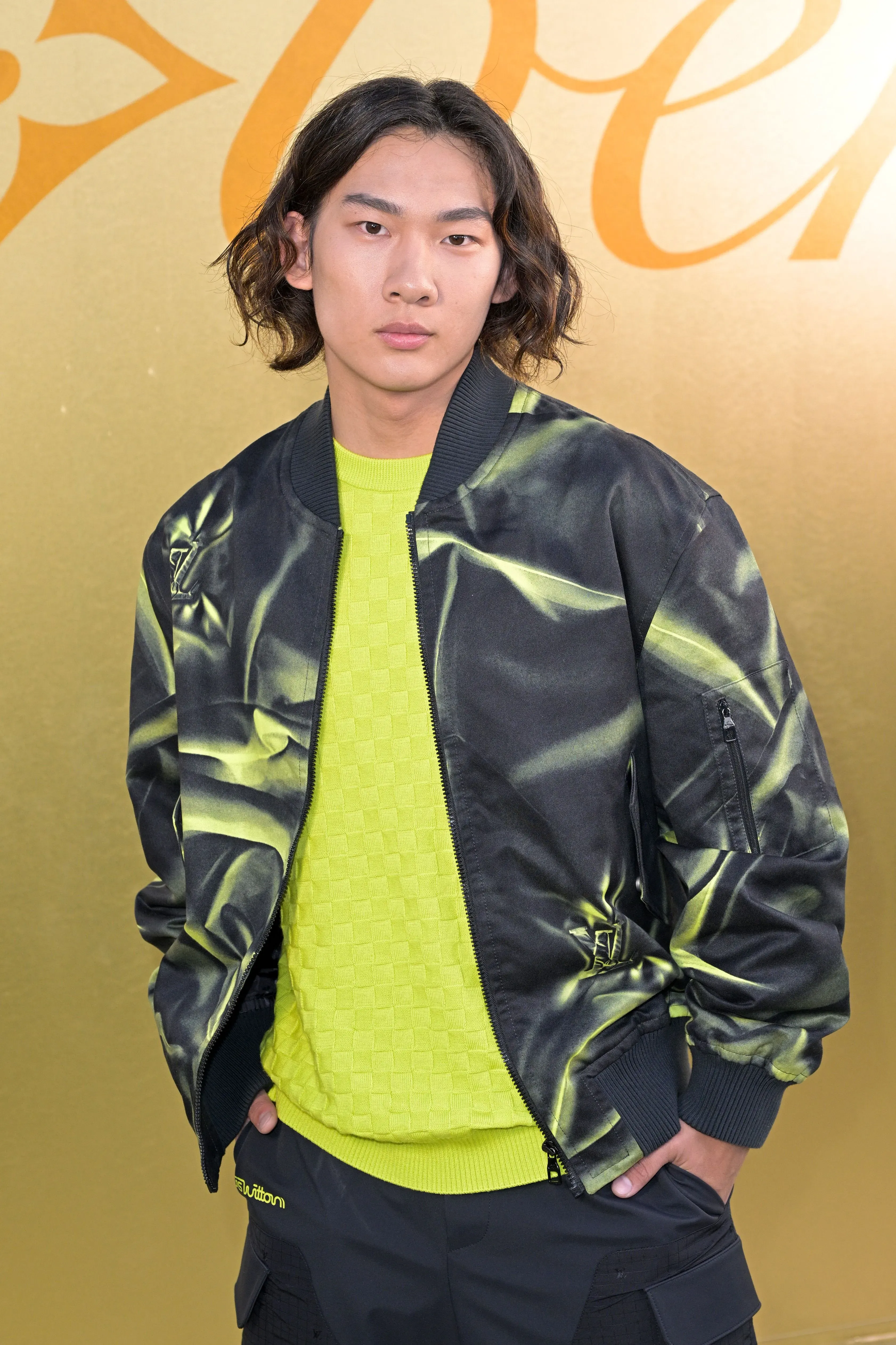 سو ييمينج، المتزلج الصيني الشاب، يرتدي سترة أنيقة ويقف أمام خلفية ذهبية، معبراً عن نجاحه بعد فوزه بالميدالية الذهبية في الألعاب الأولمبية.