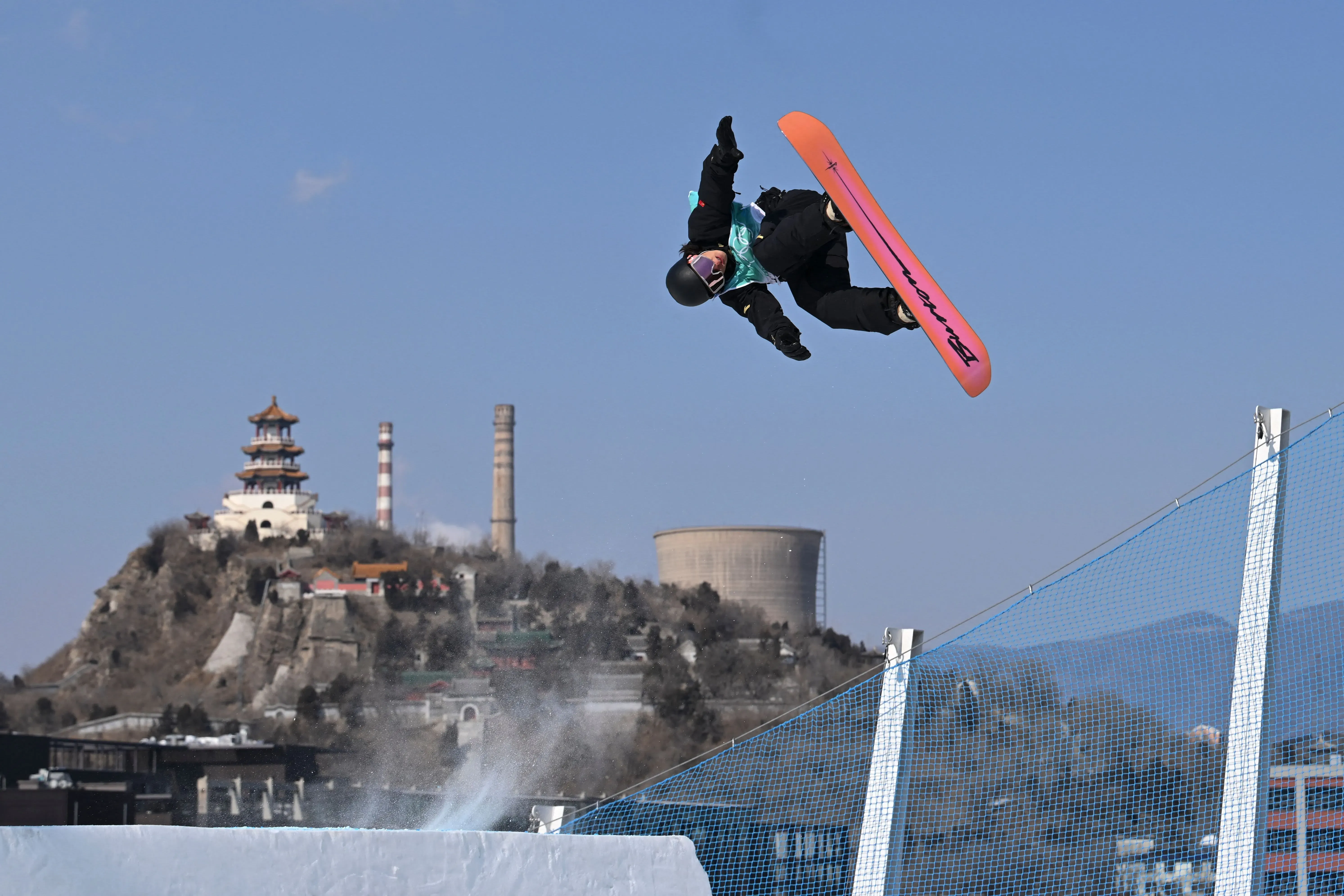 سوي ييمينغ، المتزلج الصيني، يؤدي حركة مذهلة على لوح التزلج في دورة الألعاب الأولمبية الشتوية في بكين، مع معالم خلفية بارزة.