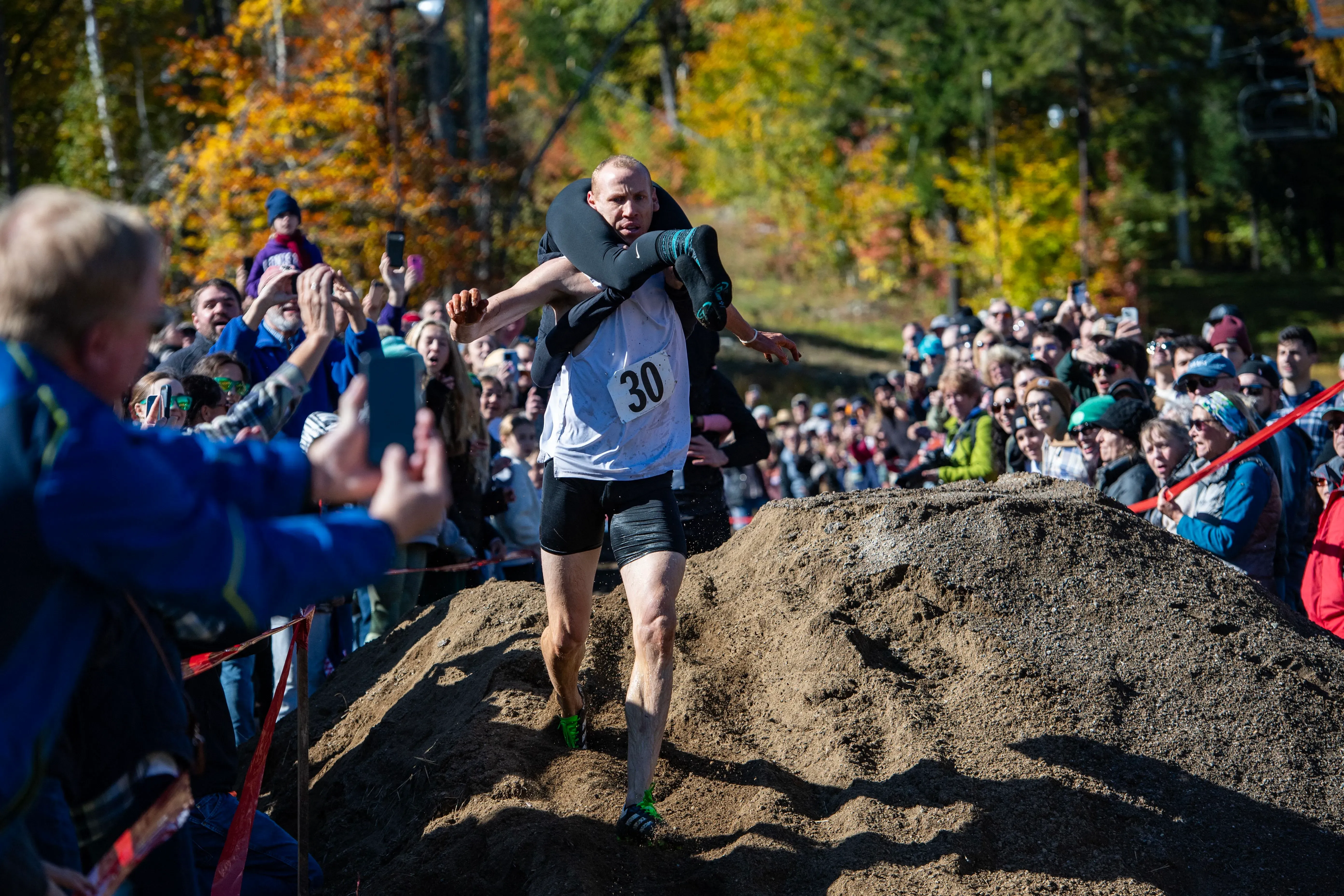 رجل يحمل زوجته في سباق عوائق، متجاوزًا حواجز في بيئة طبيعية، مع جمهور متحمس يشاهد.