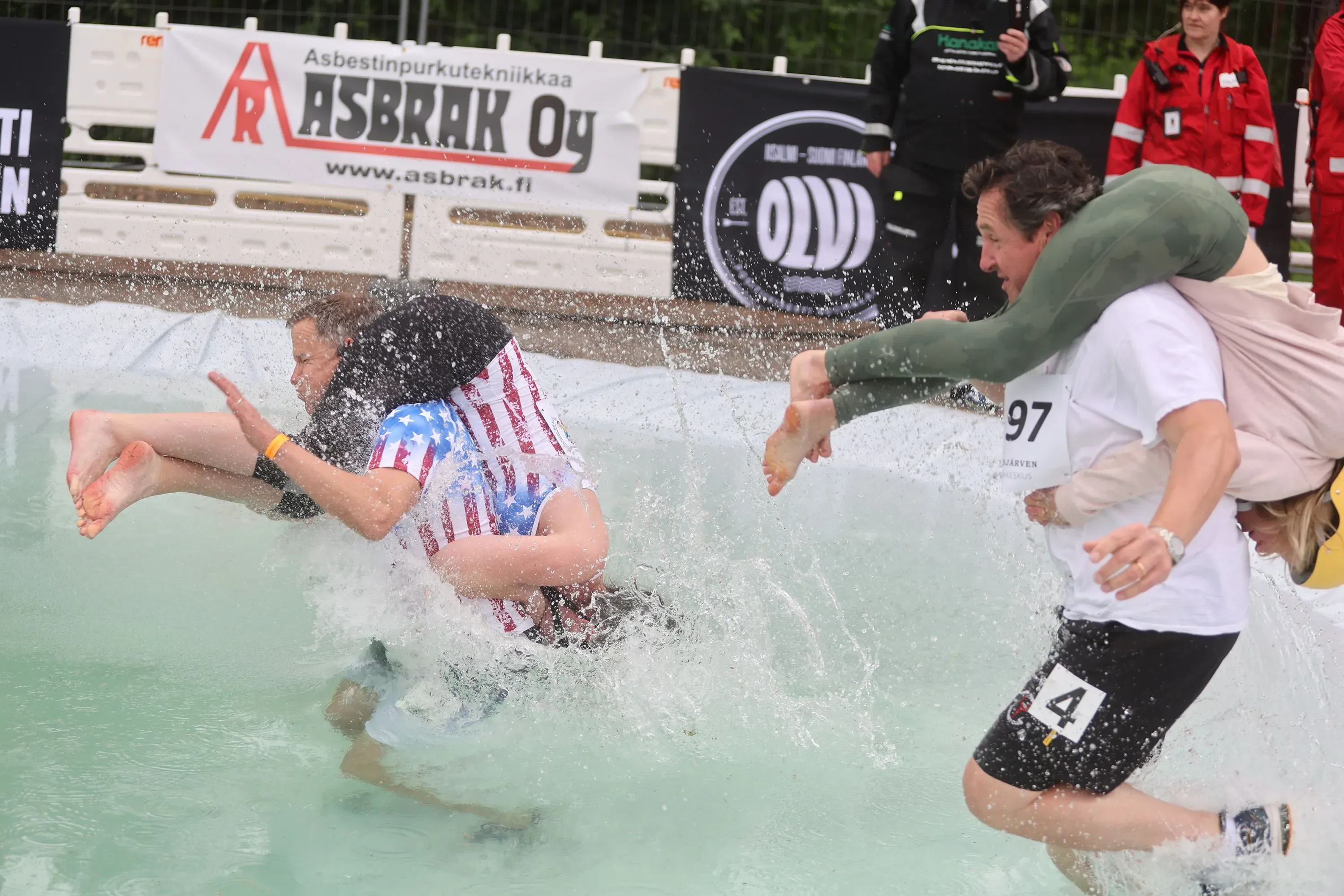 زوج يحمل زوجته أثناء سباق في الماء، يظهران في لحظة مثيرة وسط حواجز، مع لافتات خلفية تشير إلى البطولة.