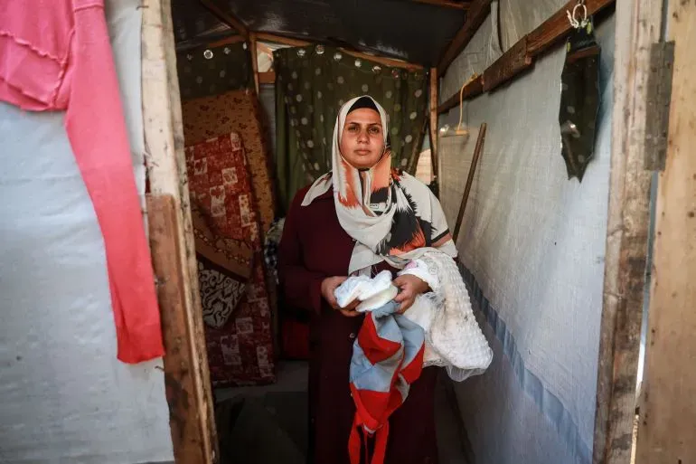 امرأة ترتدي حجابًا وتحمل ملابس مبللة في مدخل خيمة، تعكس معاناة العائلات النازحة في دير البلح جراء الأمطار الغزيرة.