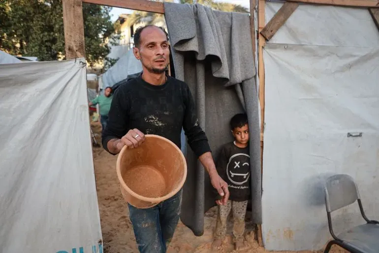 رجل يحمل دلوًا في مخيم للنازحين بدير البلح، بينما يقف طفل خلفه، في مشهد يعكس معاناة العائلات بسبب الأمطار الغزيرة.