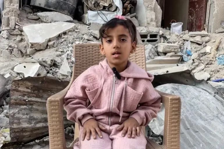 فتاة صغيرة تجلس على كرسي في منطقة مدمرة بغزة، تعبر عن مشاعرها بعد فقدان والديها جراء القصف، وتظهر آثار الحزن على وجهها.
