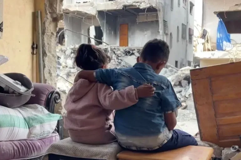 طفلان يجلسان معًا في منزل مدمر بغزة، يراقبان الدمار من النافذة، معبرين عن الحزن والارتباط في ظل الظروف الصعبة.