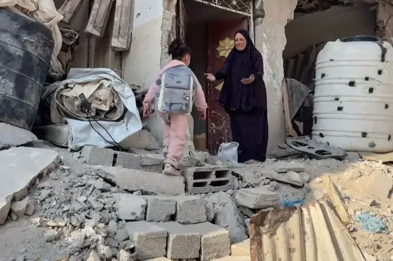 طفلة صغيرة تحمل حقيبة مدرسية تتجه نحو منزل مدمر في غزة، بينما امرأة ترتدي عباءة تقف على عتبة الباب.
