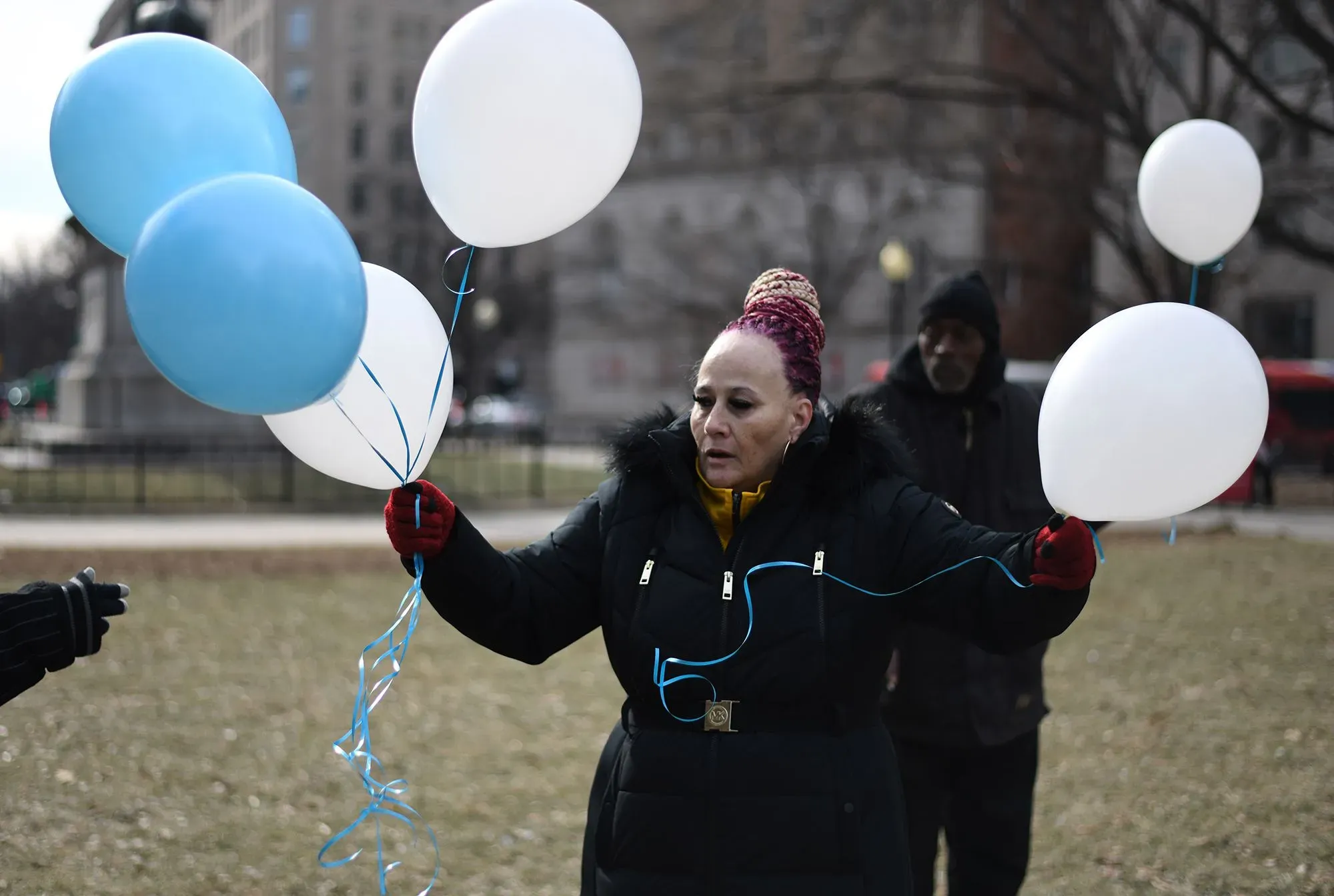 امرأة تحمل بالونات زرقاء وبيضاء في حديقة، تعبيرها يظهر الأمل والتحدي، في سياق مناقشة حول برنامج الإسكان أولاً لمساعدة المشردين.