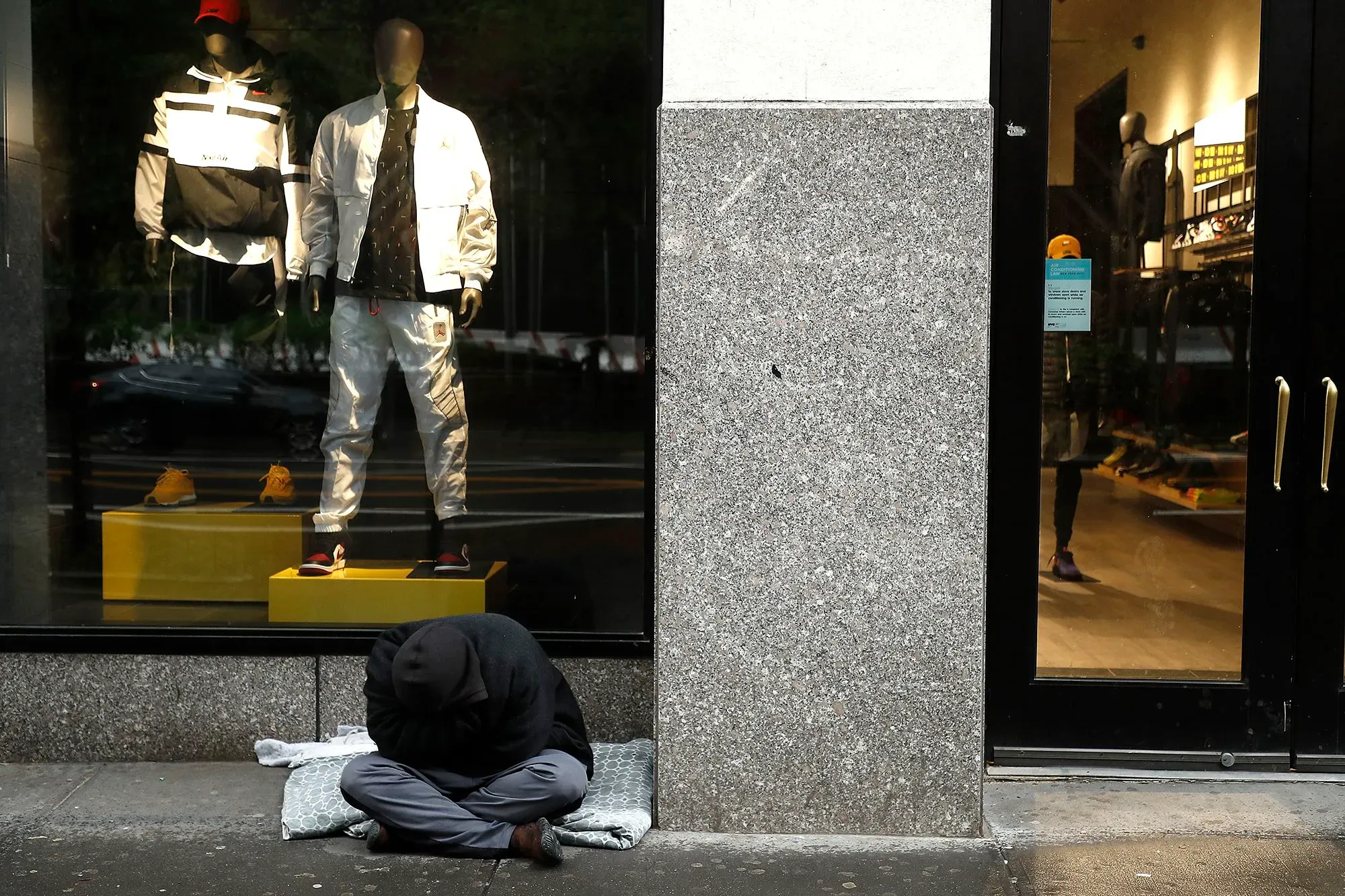 امرأة جالسة على الرصيف، تغطي رأسها، بجوار واجهة متجر تعرض ملابس رياضية جديدة، تعكس تحديات التشرد في واشنطن.