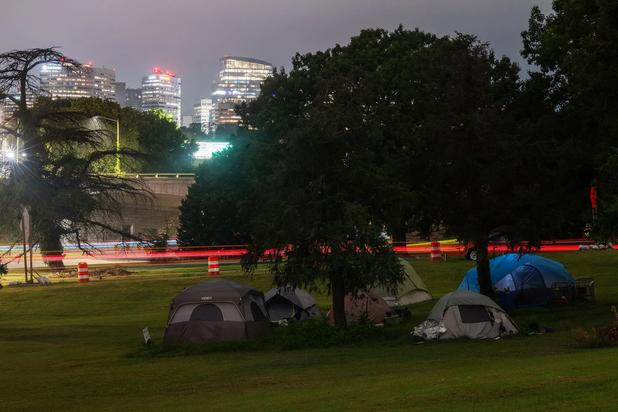 تظهر الصورة مجموعة من الخيام في حديقة وسط المدينة، مع خلفية لمباني عالية مضاءة، مما يعكس واقع التشرد في المناطق الحضرية.