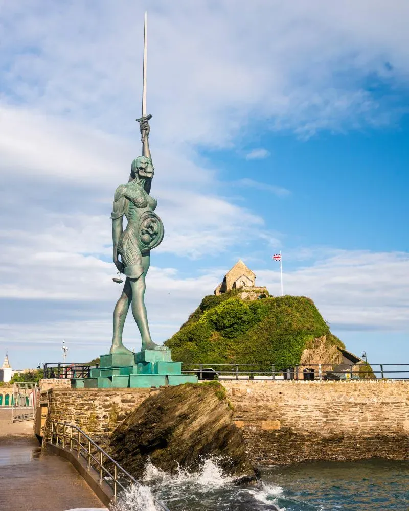 تمثال برونزي لجندي يحمل سيفًا عالياً، يقف على صخرة بجوار البحر، مع خلفية لجزيرة صغيرة وقلعة، تحت سماء زرقاء.