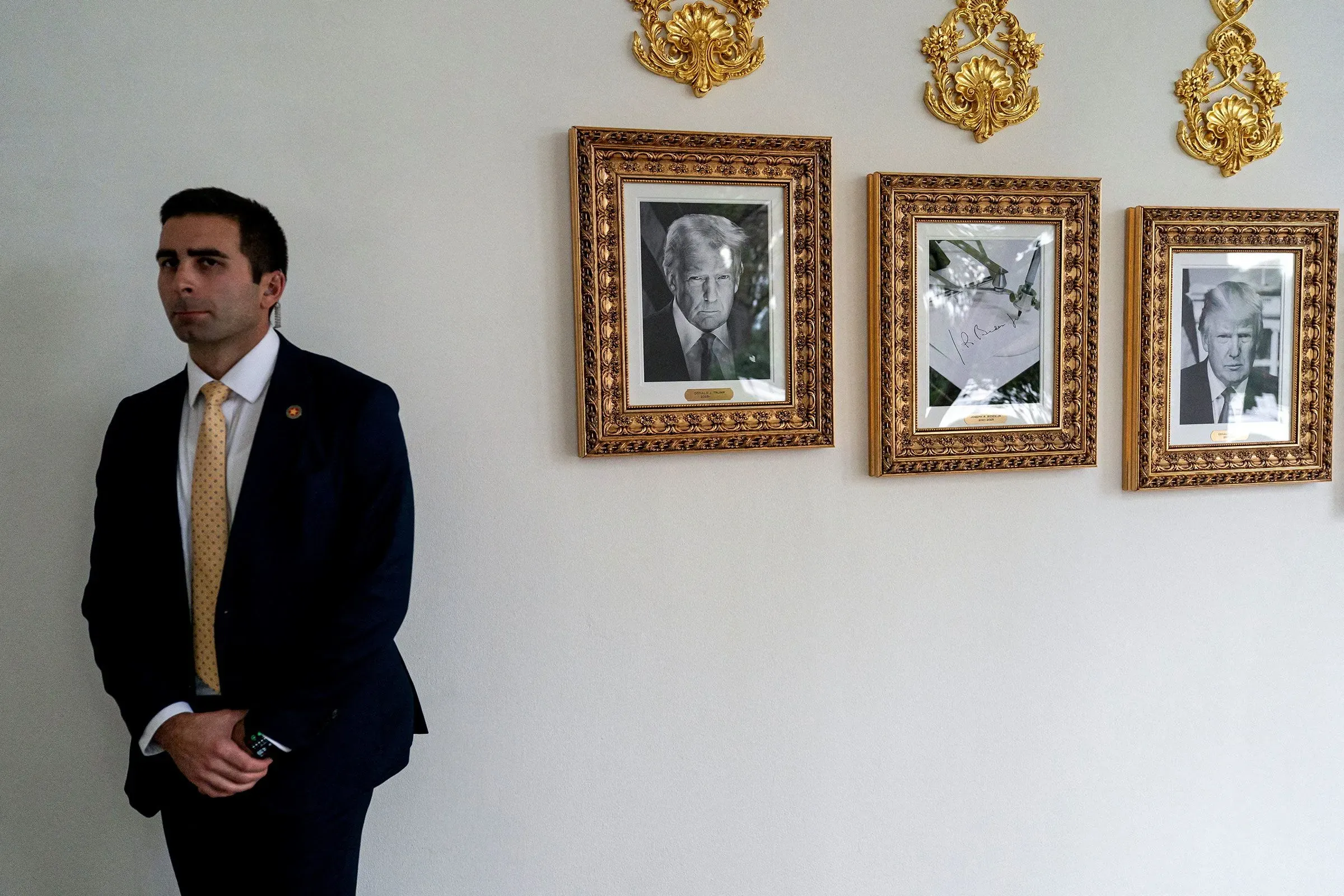 شخص واقف بجانب جدار يحمل صورًا مؤطرة لترامب، مع تعبير جاد، يعكس أجواء سياسية مشحونة.