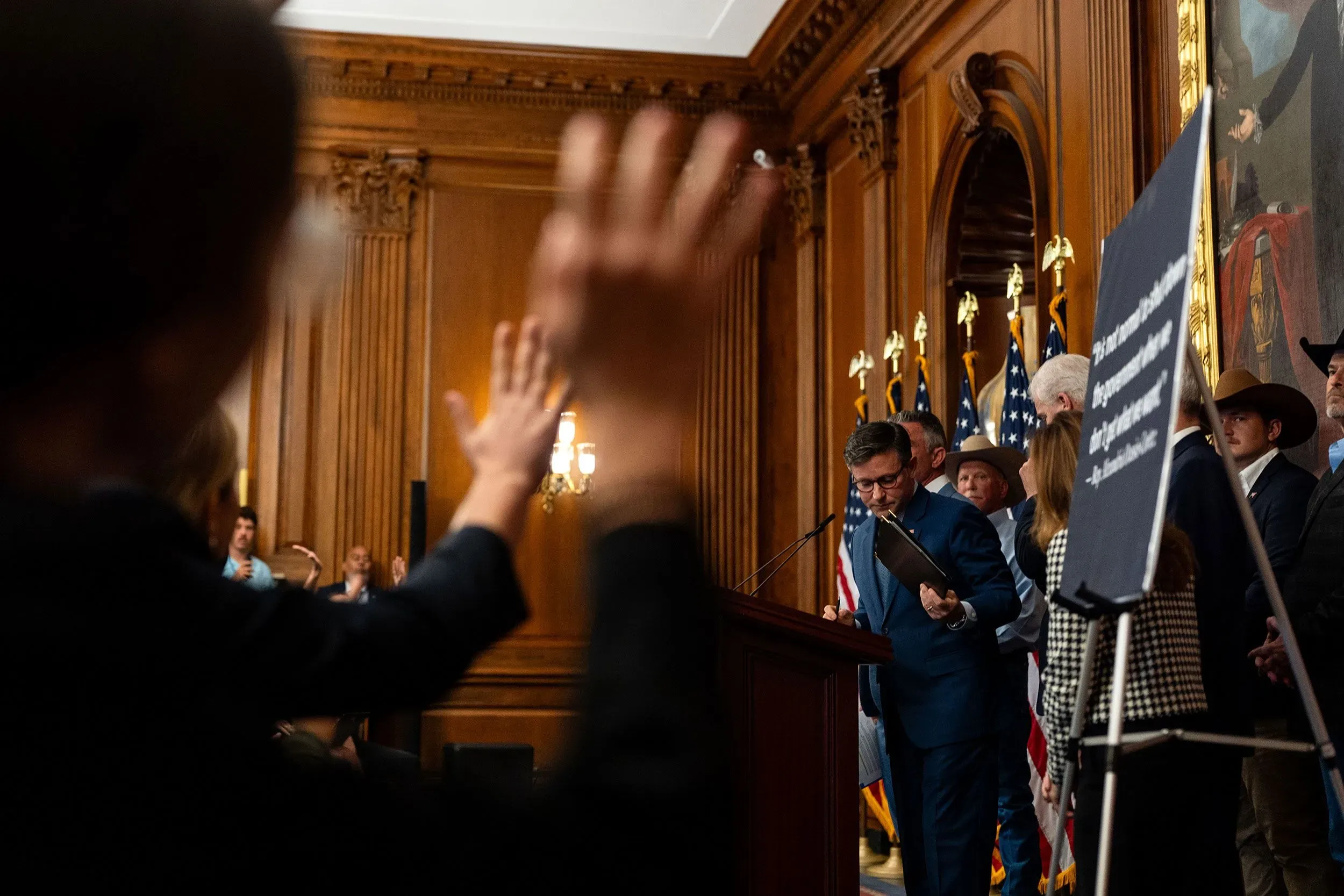 مؤتمر صحفي في مبنى حكومي، حيث يتحدث متحدث رسمي مع وجود حشد من الناس في الخلفية، مع لافتات تحمل رسائل سياسية.