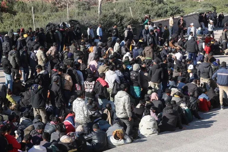 مجموعة كبيرة من طالبي اللجوء يجلسون في منطقة مفتوحة، يرتدون ملابس شتوية، بعد إنقاذهم في كريت.