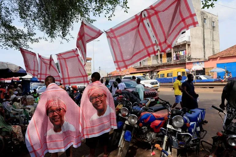 شخصان يرتديان أقمشة تحمل صورة الرئيس أومارو سيسوكو إمبالو في سوق مزدحم، مع وجود لافتات تتطاير في الهواء.