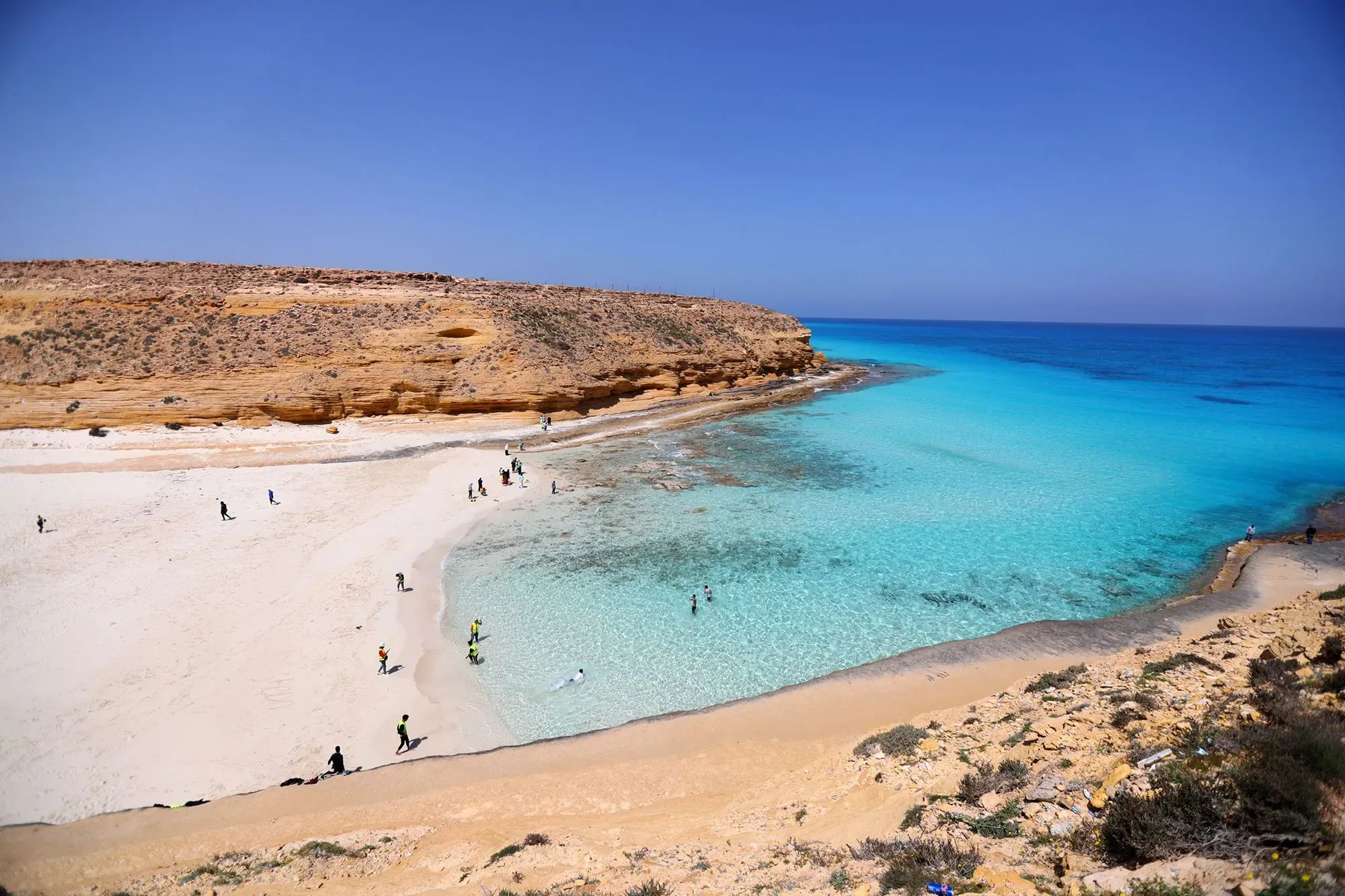 شاطئ ساحل مصر الشمالي يظهر فيه زوار يستمتعون بالمياه الزرقاء الصافية والرمال البيضاء، محاطًا بتضاريس طبيعية.