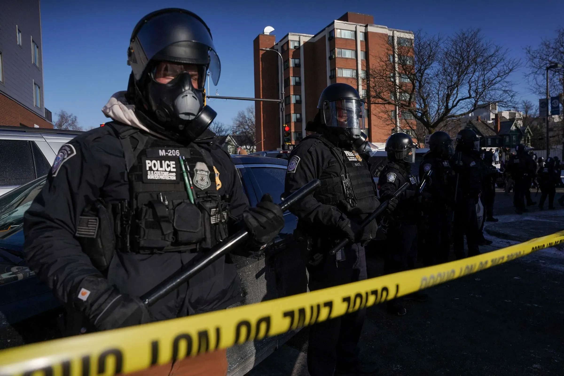 رجال شرطة يرتدون دروعًا وخوذًا واقية يقفون خلف شريط تحذيري في موقع حادث مينيابوليس، مما يعكس التوترات المتعلقة بقضايا السلاح وحقوق الأفراد.