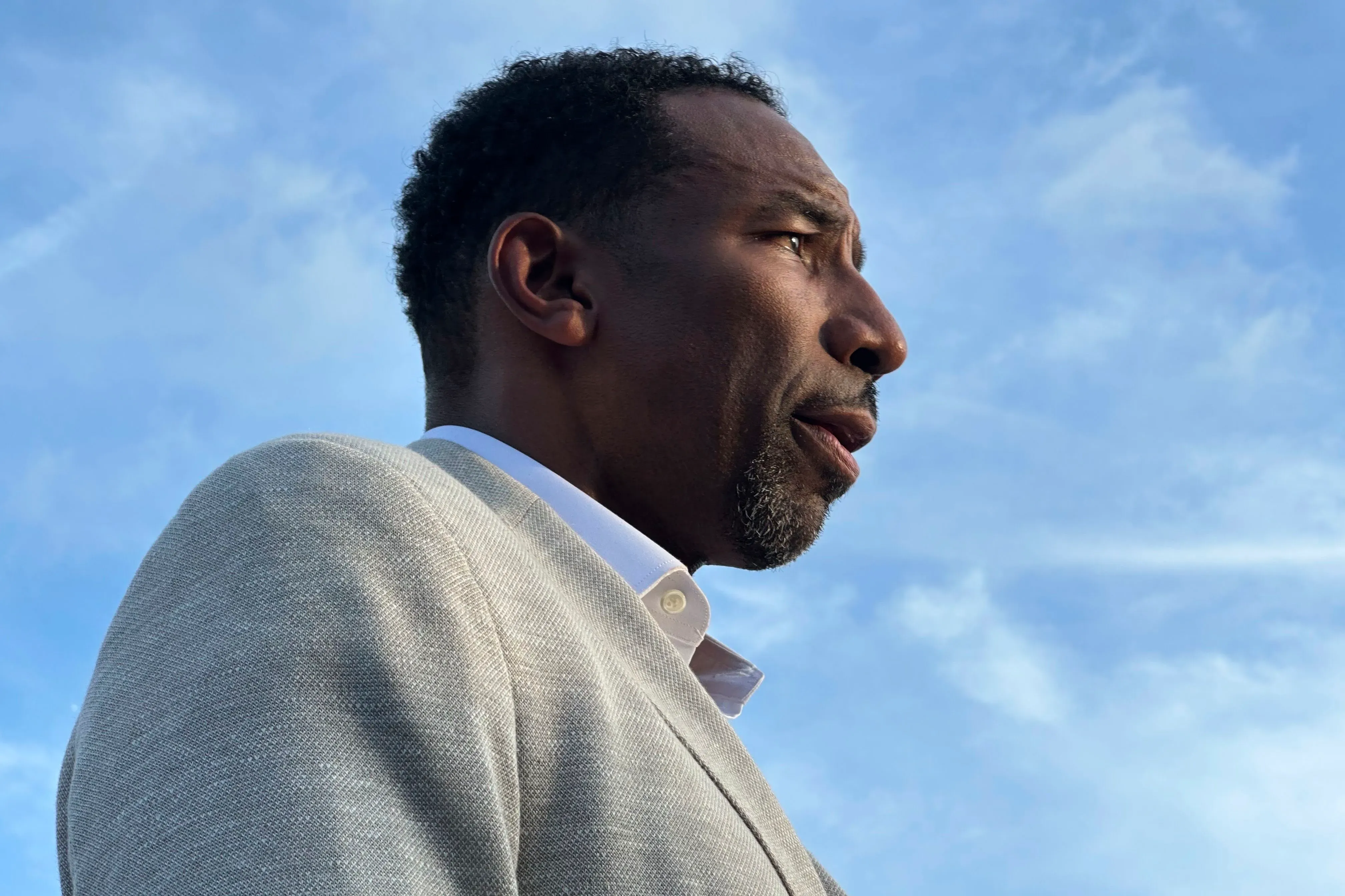 صورة لرجل يرتدي بدلة رمادية، ينظر إلى الأفق تحت سماء زرقاء. تعكس تعابير وجهه الجدية والتفكير.