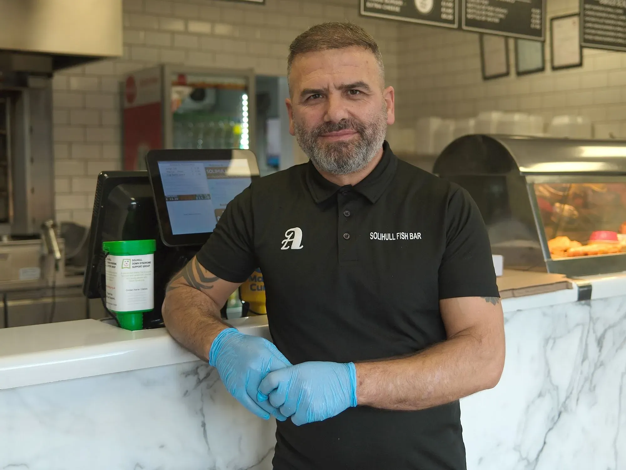 رجل يقف في متجر السمك والبطاطس المقلية في سوليهول، يرتدي قفازات زرقاء، مع خلفية تحتوي على قائمة الطعام وأدوات الطهي.