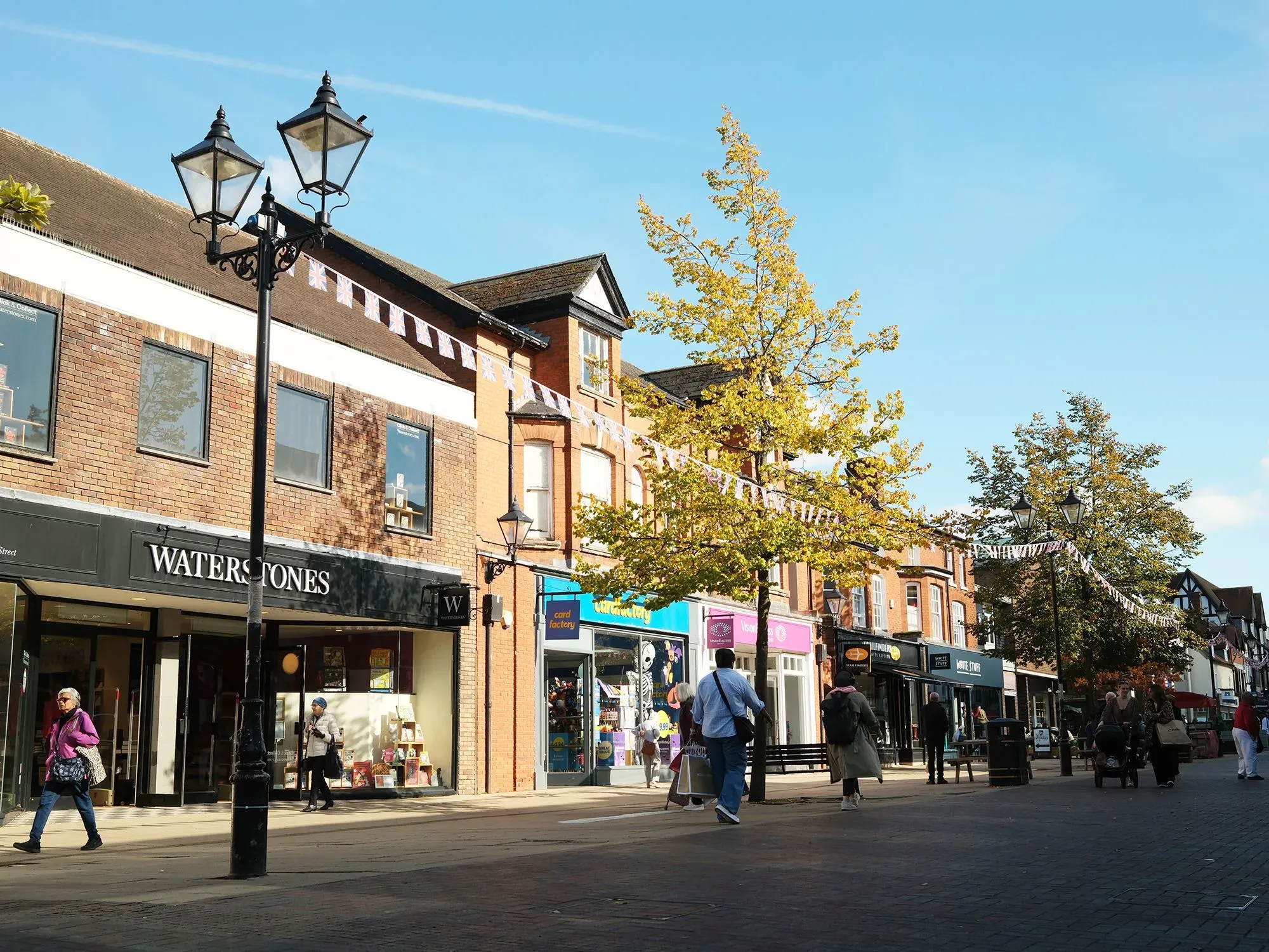 شارع مزدحم في وسط مدينة سوليهول، مع متاجر متنوعة وأشجار تتلون بألوان الخريف، مما يعكس حيوية الحياة التجارية بعد جائحة كورونا.