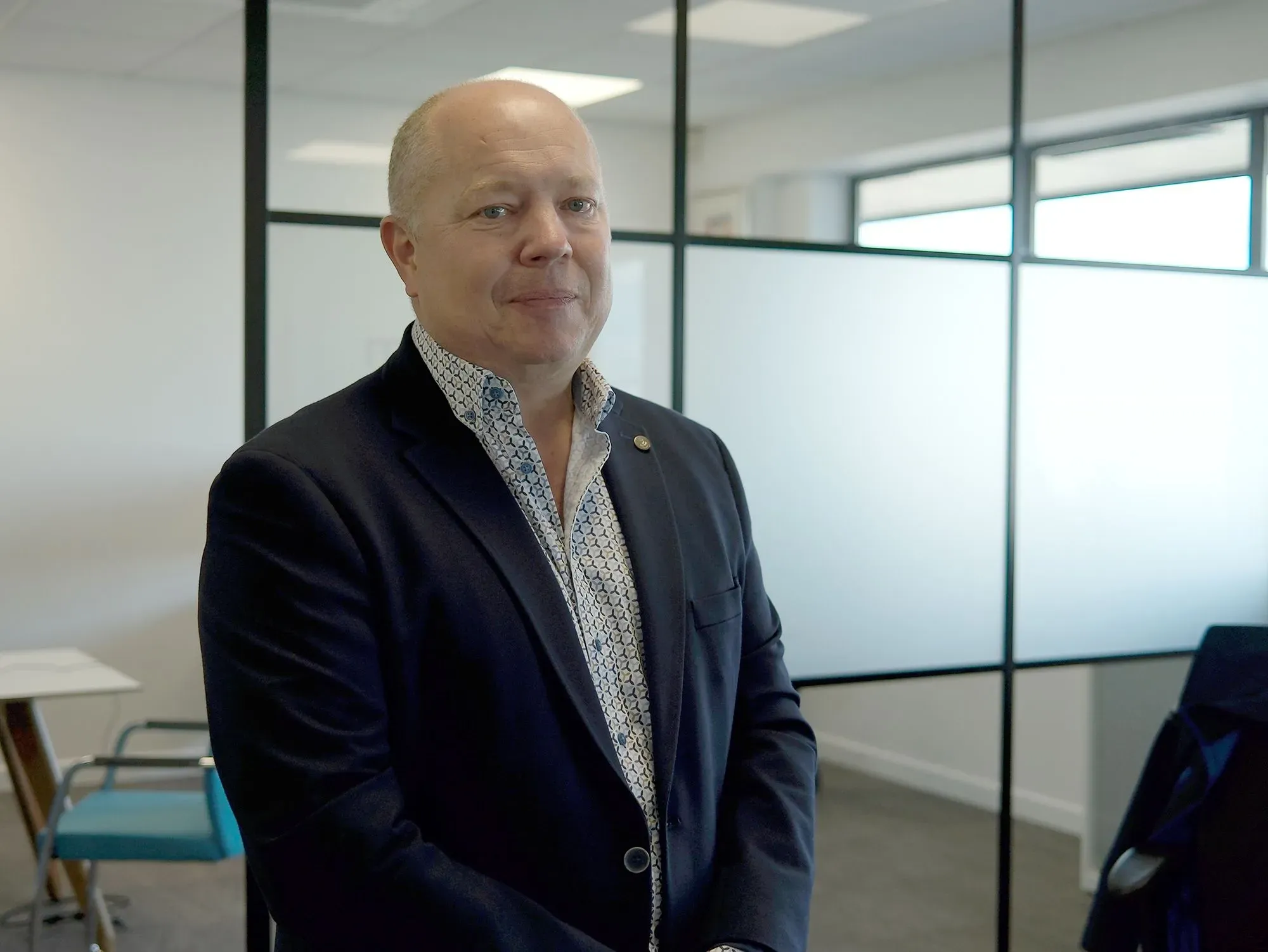 رجل في بدلة أنيقة يقف مبتسمًا في مكتب حديث، مع جدران زجاجية تفصل بين المساحات. تعكس الصورة أجواء العمل المهنية.