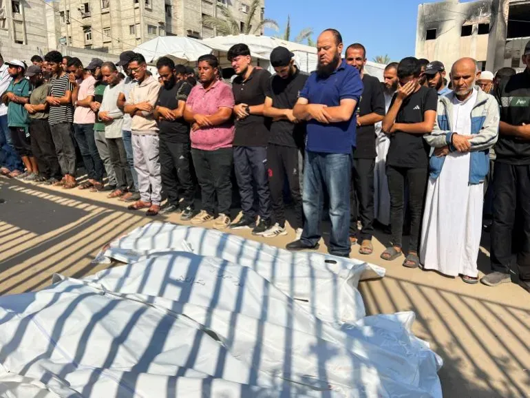 مجموعة من الأشخاص يرتدون ملابس سوداء، يقفون في صف أثناء تأدية الصلاة على جثامين ضحايا العدوان في غزة، مع خلفية لمبانٍ مدمرة.