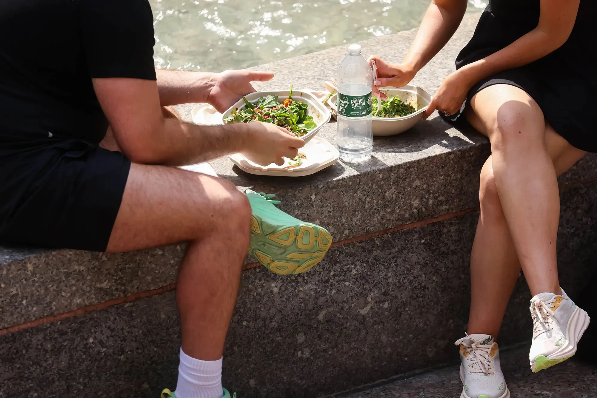 شخصان يجلسان على حافة مائية، يتناولان سلطات صحية من أطباق قابلة للتخصيص، مع زجاجة ماء بجانبهم.
