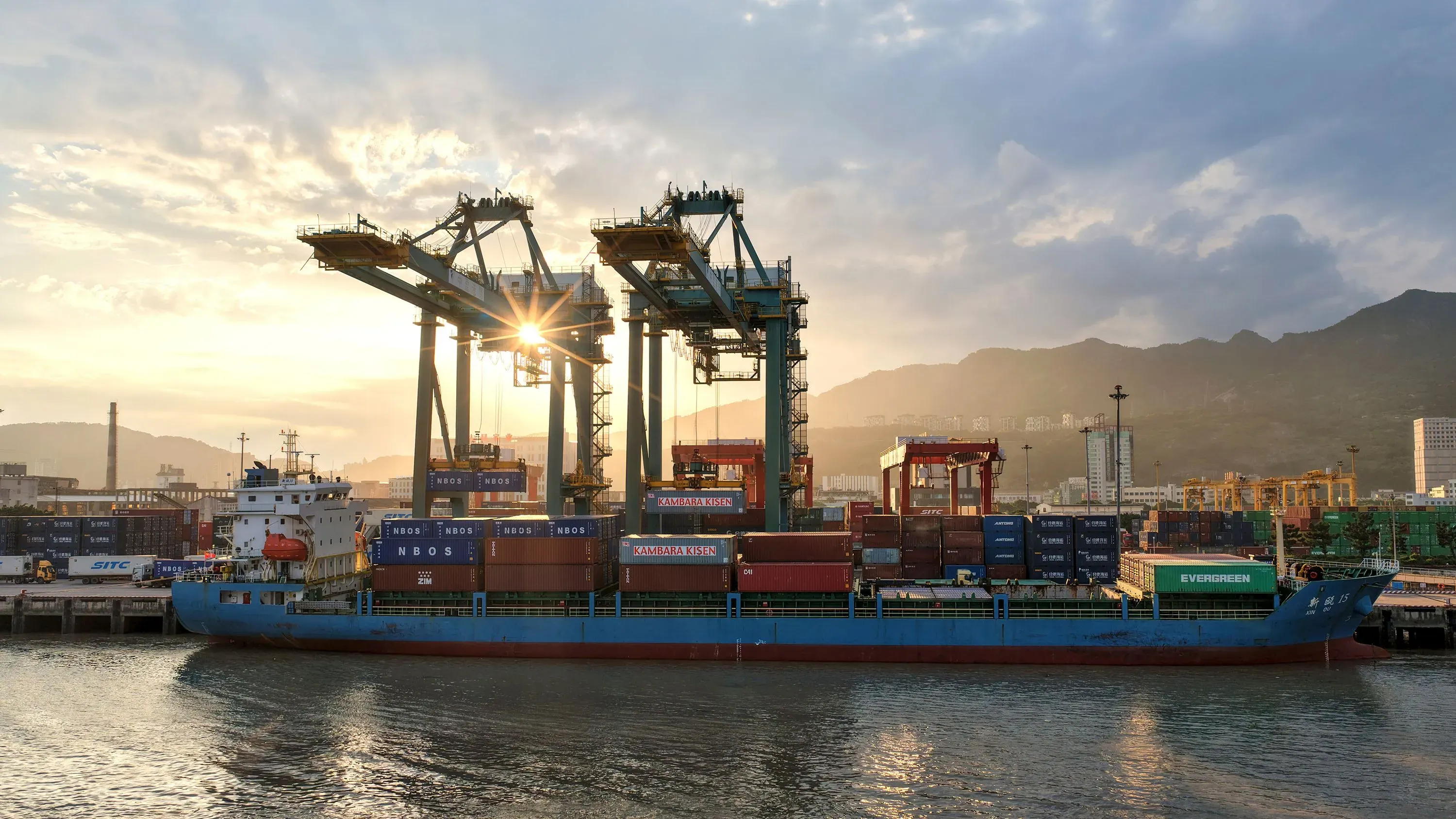 سفينة شحن محملة بالحاويات في ميناء مع وجود رافعات خلفها، تعكس أشعة الشمس في السماء، مما يرمز للتجارة العالمية والاقتصاد.