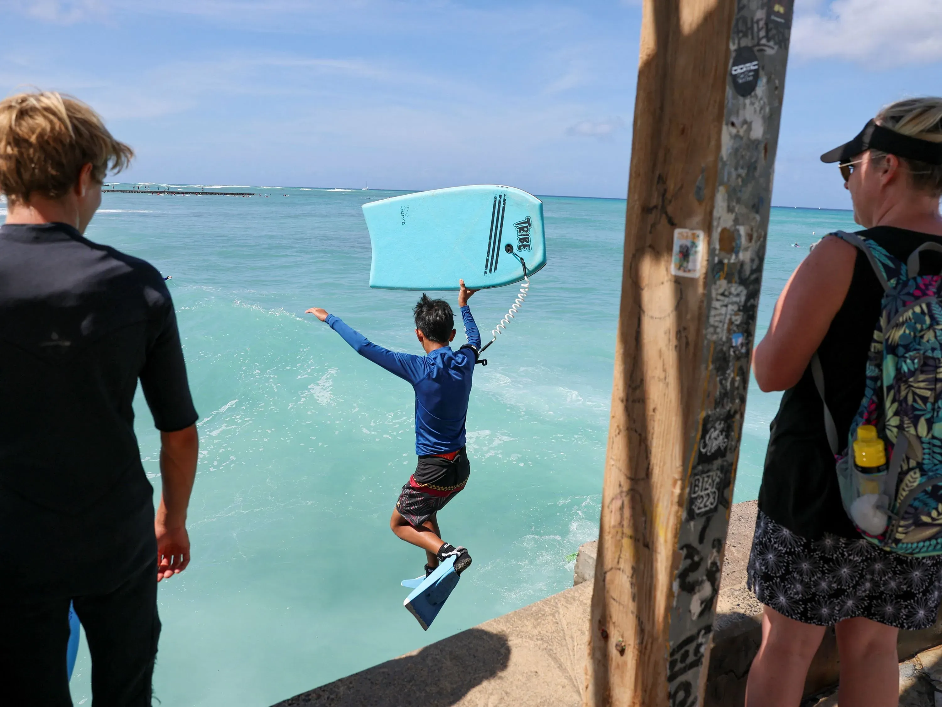 طفل يقفز إلى المياه من رصيف في وايكيكي، بينما يراقبه آخرون، مما يعكس أجواء المرح بعد تحذيرات التسونامي.