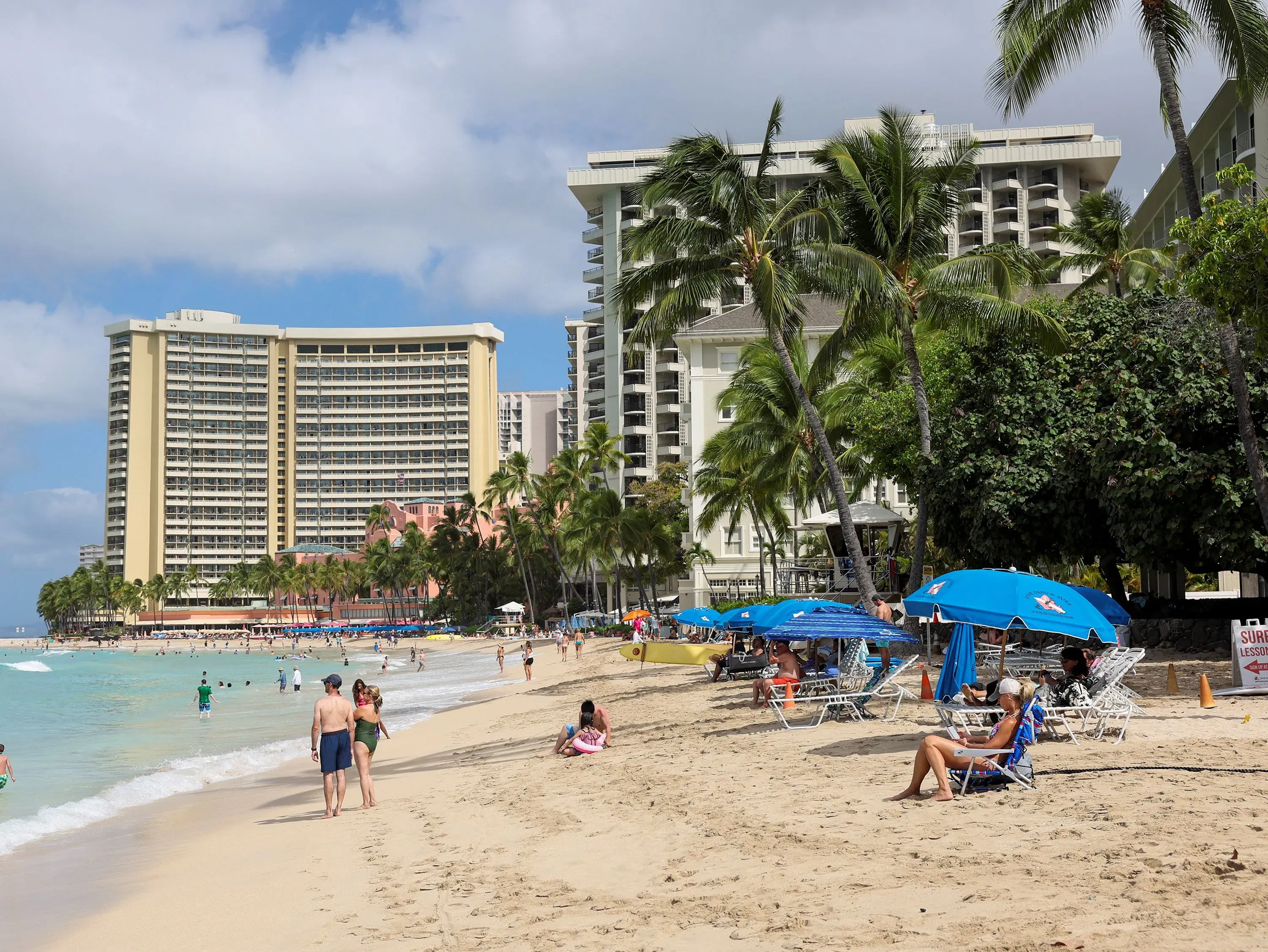 شاطئ وايكيكي في هاواي مزدحم بالسياح، حيث يستمتع الناس بالماء والشمس، مع وجود فنادق وأشجار النخيل في الخلفية.