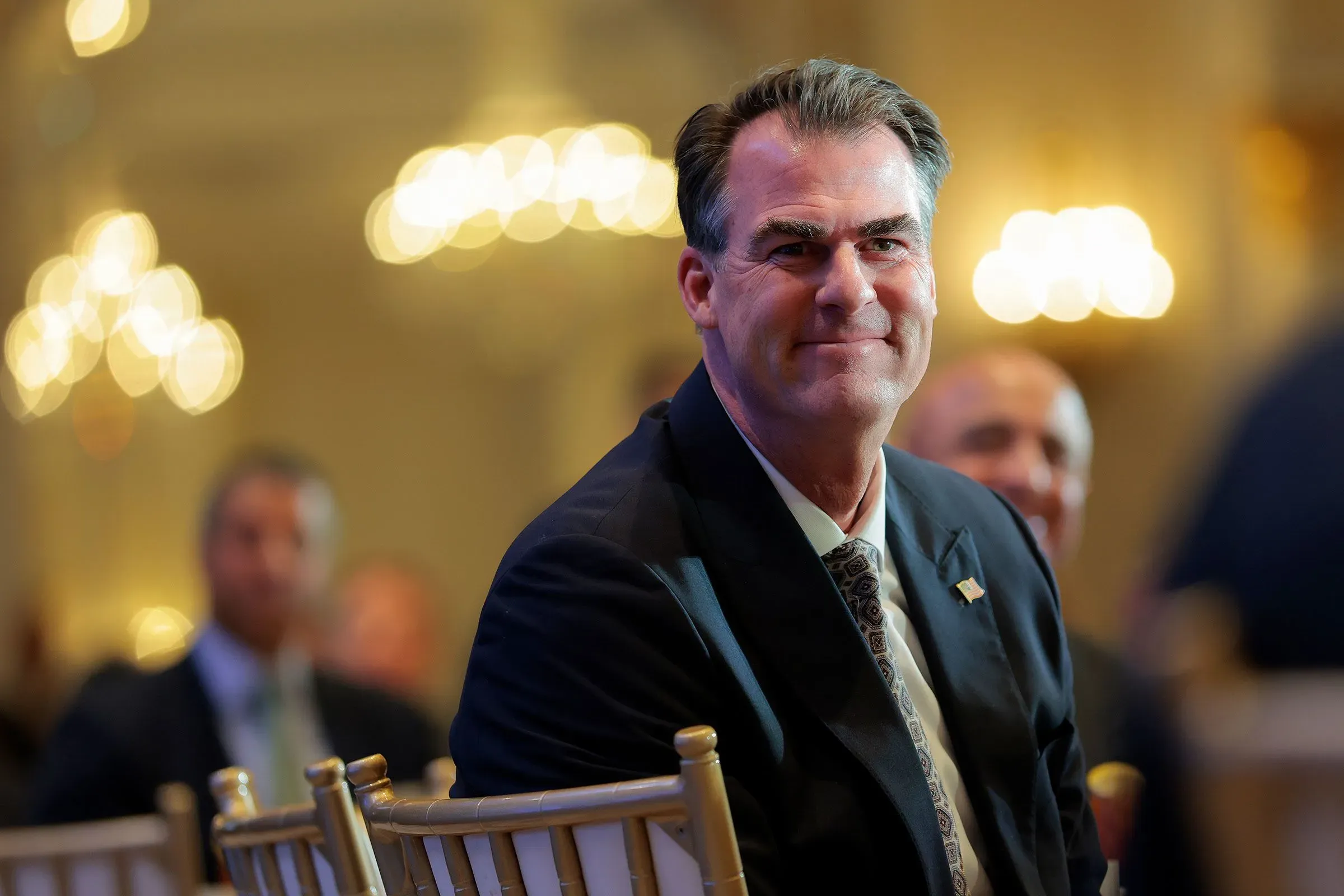 حاكم ولاية أوكلاهوما كيفن ستيت مبتسم خلال حدث رسمي، مع أضواء خافتة في الخلفية، مما يعكس التوترات السياسية مع ترامب.