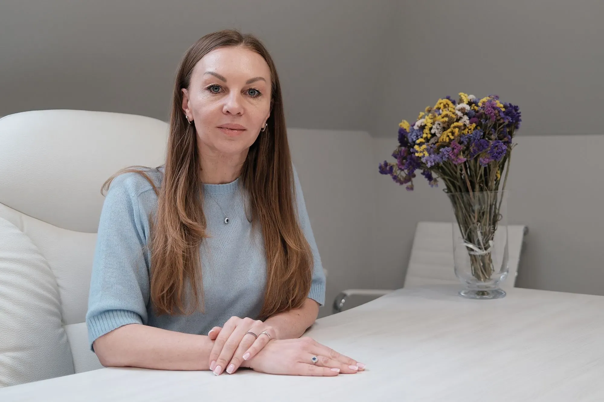 امرأة تجلس على طاولة، ترتدي سترة زرقاء، مع باقة من الزهور الملونة خلفها. تعكس ملامحها مشاعر القلق والتفكير في قضايا العنف الأسري في أوكرانيا.