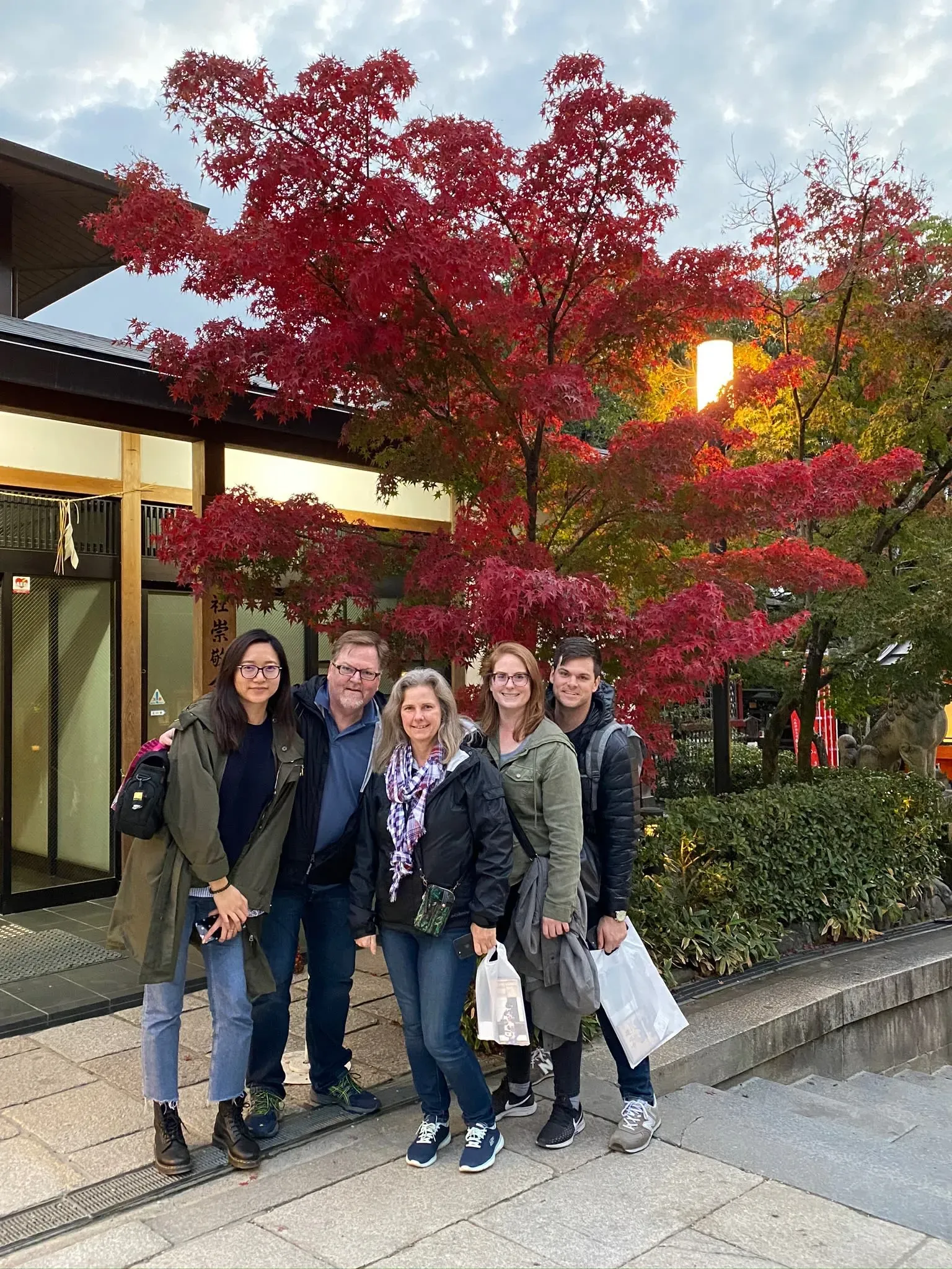 مجموعة من خمسة أشخاص يقفون معاً أمام شجرة ذات أوراق حمراء، في موقع جميل، مع مبنى خلفهم وتفاصيل طبيعية حولهم.