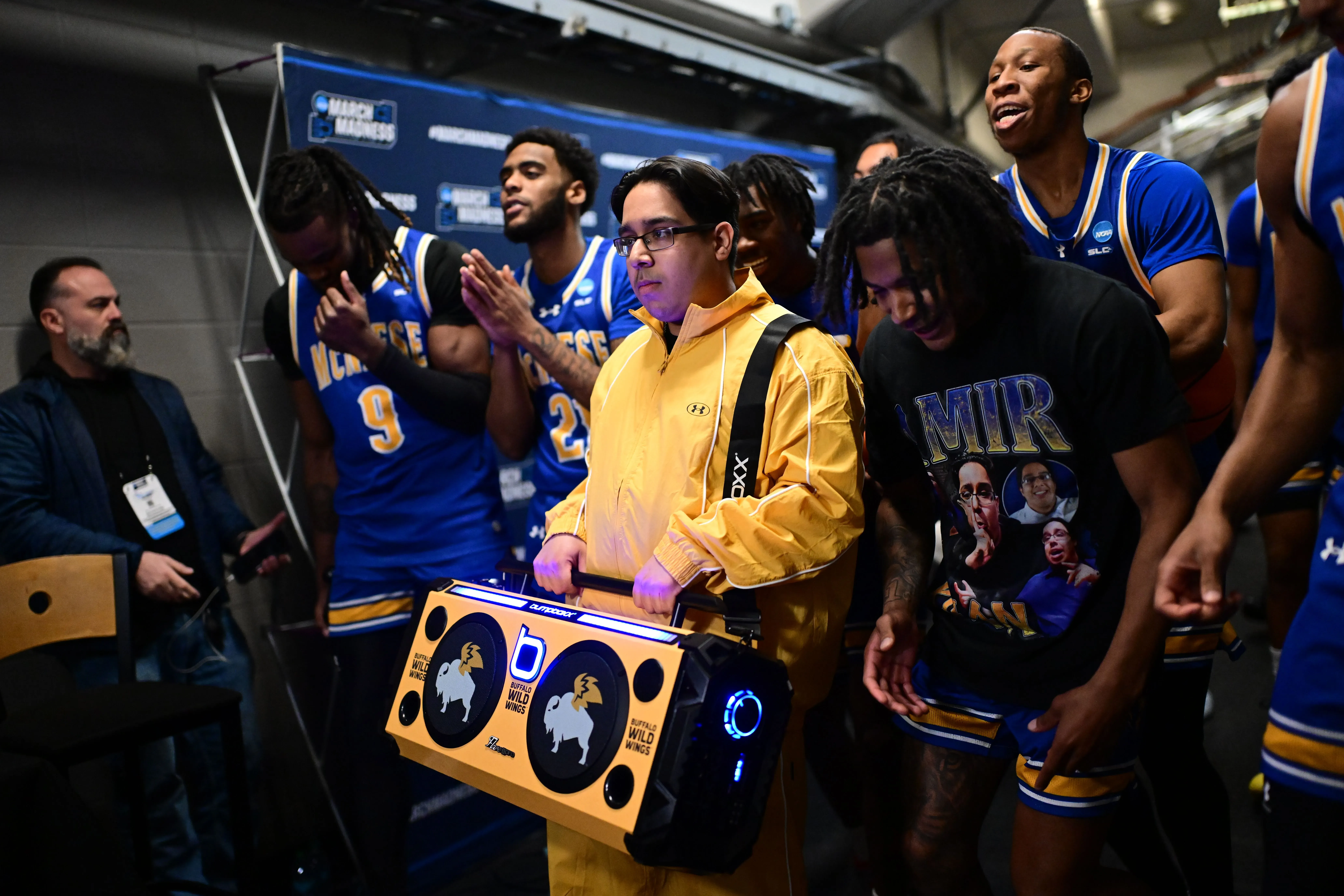 شخص يرتدي سترة صفراء يحمل جهاز تسجيل كبير أثناء دخول فريق كرة السلة ماكنيس إلى الملعب، مع وجود لاعبين آخرين خلفه.