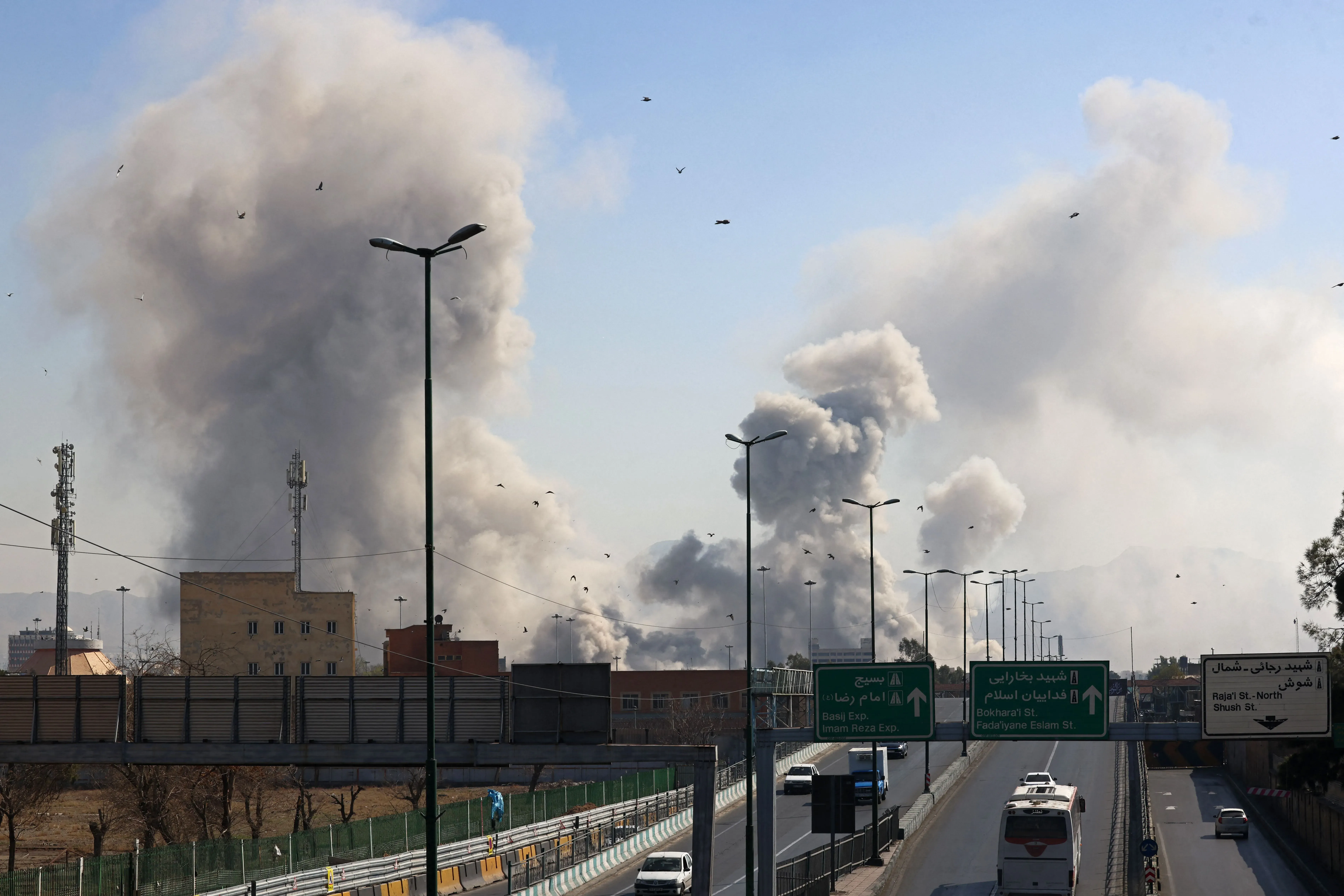 دخان كثيف يتصاعد من مواقع القصف في إيران، مع وجود لافتات طرق وإشارات مرورية في المقدمة، مما يعكس تصاعد التوترات العسكرية.