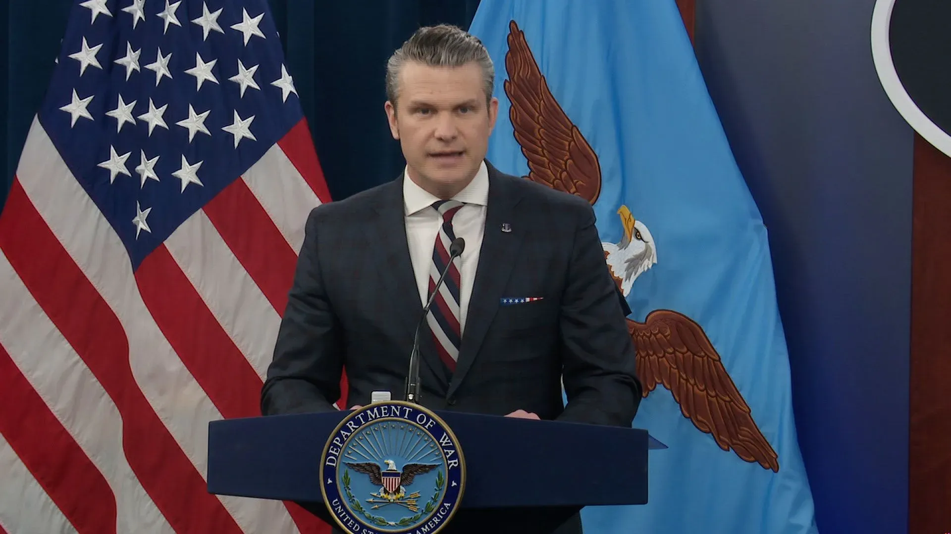 يتحدث المتحدث باسم وزارة الدفاع الأمريكية عن تكلفة الحرب مع إيران، مع وجود علمين أمريكي وإيراني خلفه.
