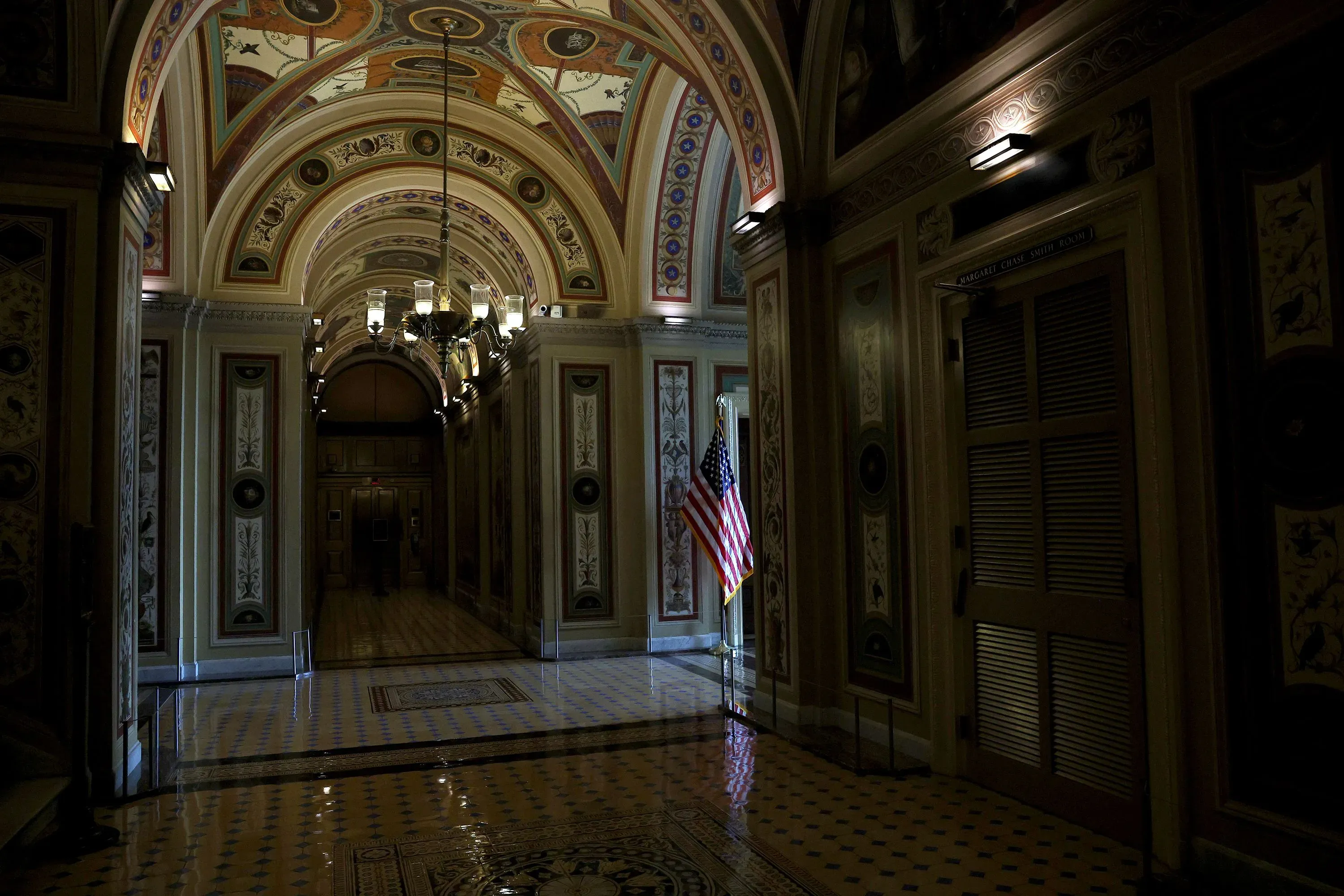 ممر مظلم في مبنى الكونغرس الأمريكي، مزين بسقف مزخرف، مع وجود علم الولايات المتحدة، مما يعكس التوترات السياسية الحالية.