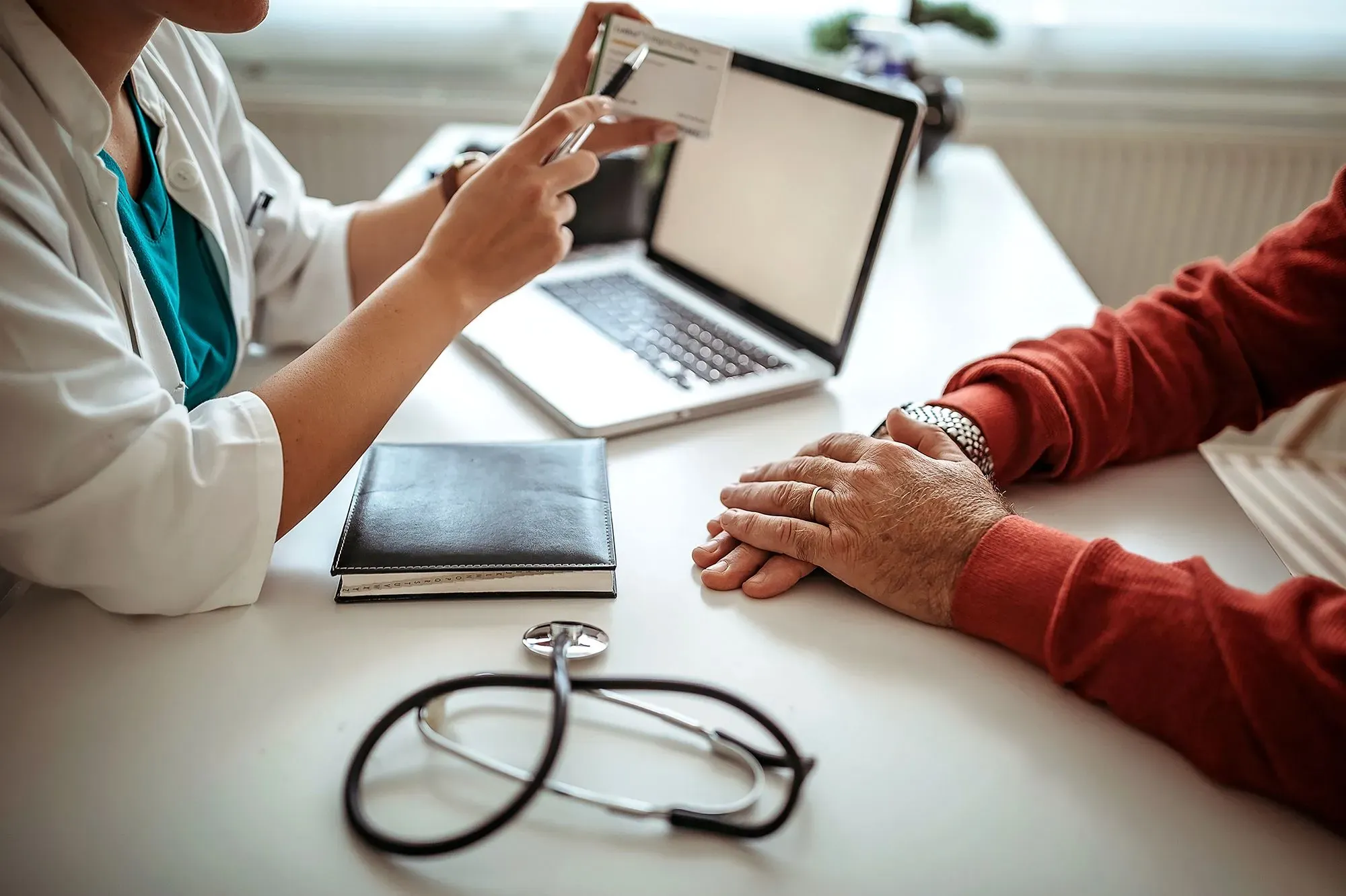 مريض يتلقى استشارة طبية، حيث يظهر طبيب يشرح تفاصيل التأمين الصحي بينما يُظهر بطاقة التأمين، مع جهاز كمبيوتر محمول على الطاولة.