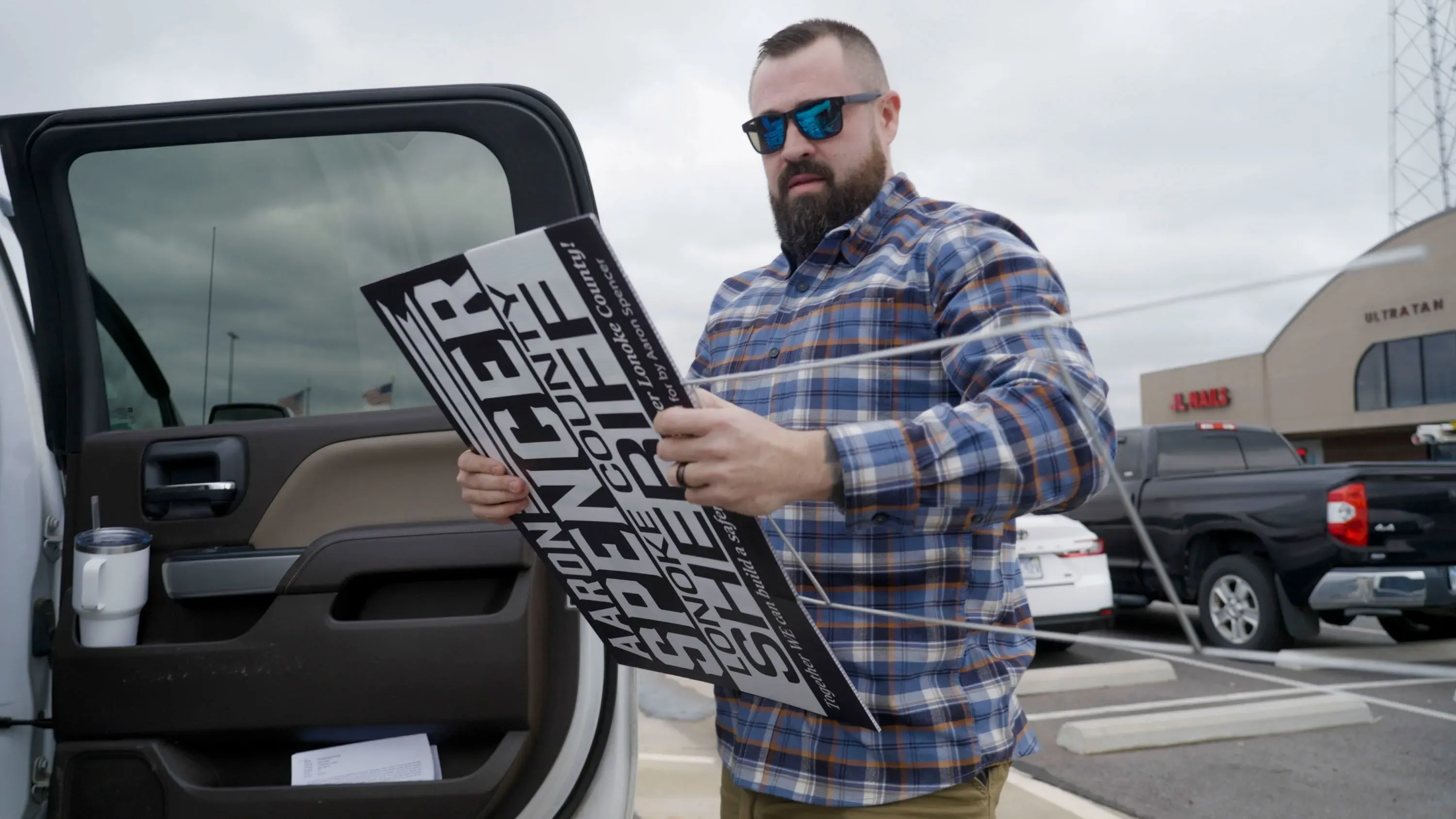 آرون سبنسر، مرشح عمدة مقاطعة لونوك، يحمل لافتة حملته الانتخابية في موقف سيارات، محاطًا بمؤيديه، تحت سماء ملبدة بالغيوم.