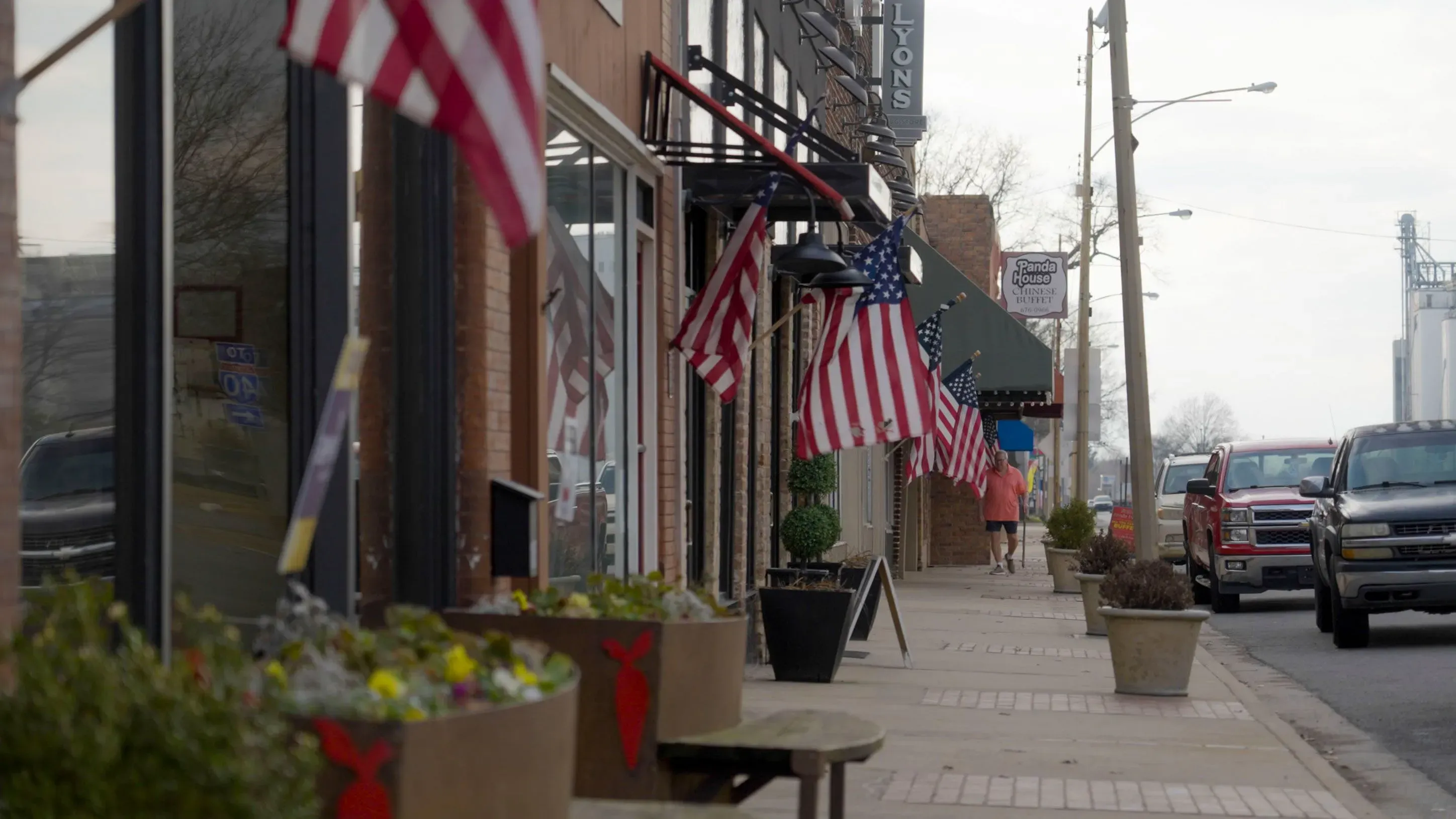 شارع تجاري في كابوت، أركنساس، مزين بأعلام أمريكية، حيث يمر الناس بجانب المحلات والمقاهي في يوم غائم.