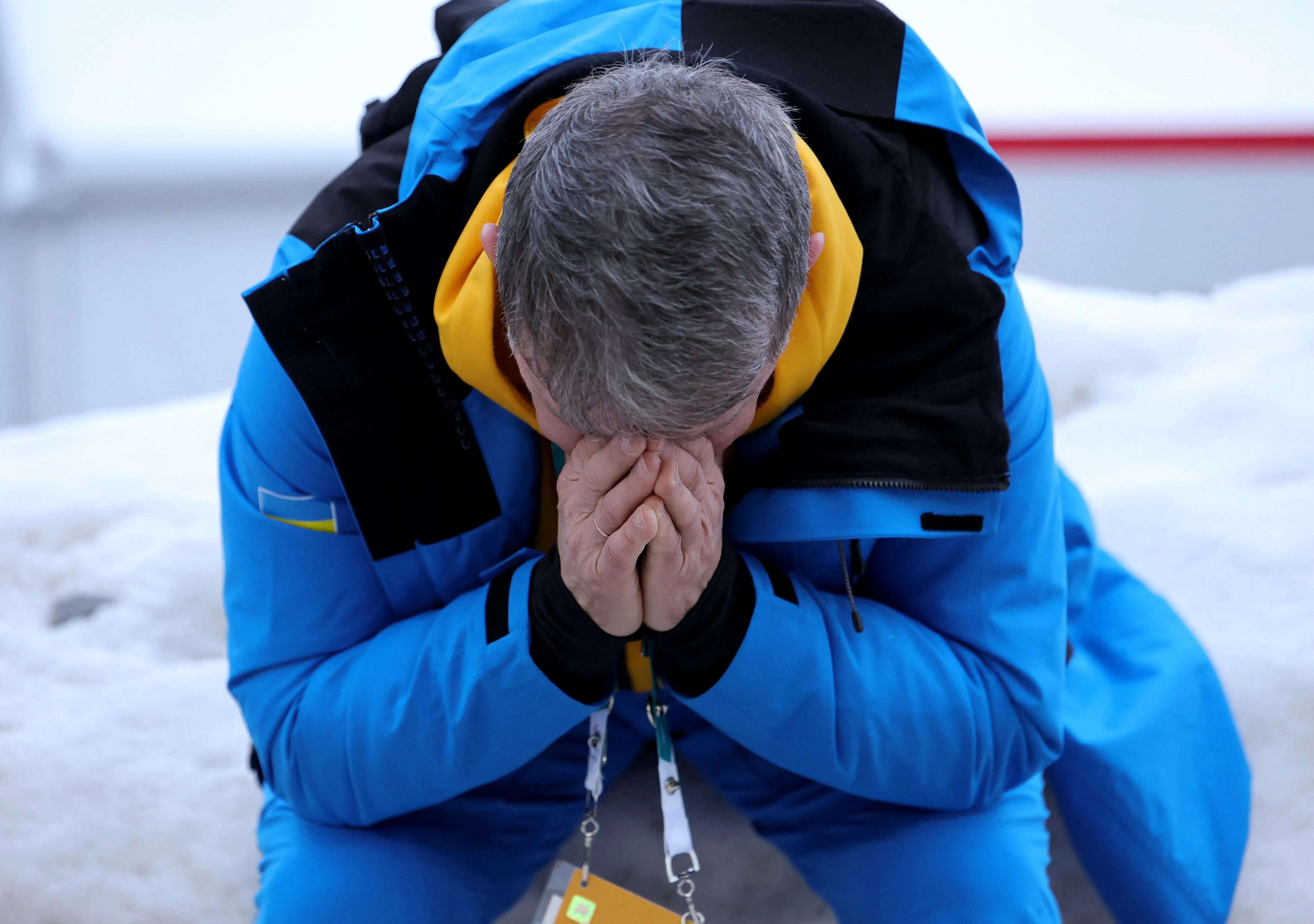 رجل أوكراني يرتدي زيًا رياضيًا أزرق، يجلس على الثلج ويضع يديه على وجهه تعبيرًا عن الحزن بعد استبعاد المتزلج هراسكيفيتش من الأولمبياد.