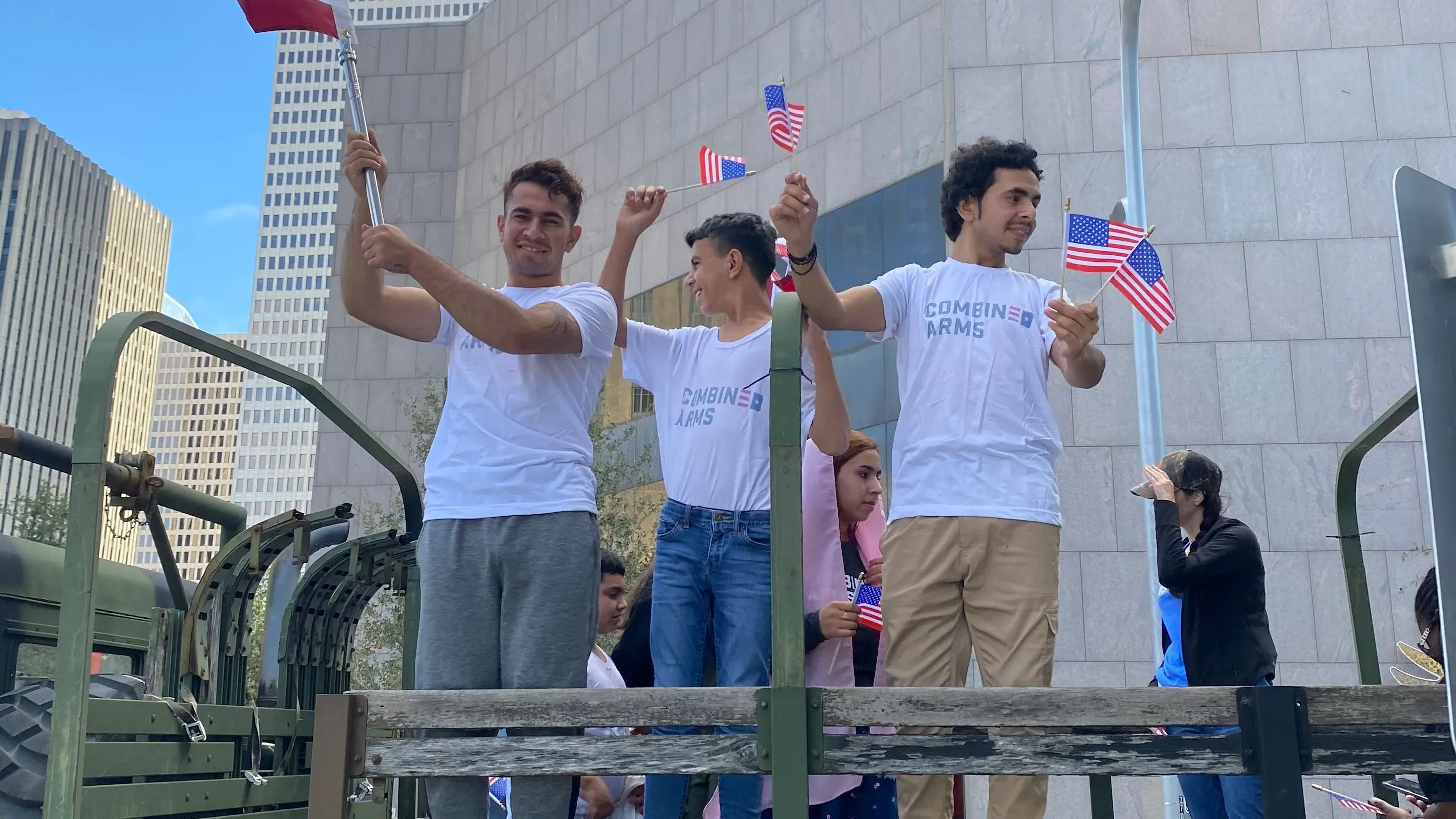 شبان يحملون أعلامًا أمريكية، يرتدون قمصان تحمل شعار "Combining Arms"، يقفون على شاحنة عسكرية خلال فعالية لدعم اللاجئين الأفغان.