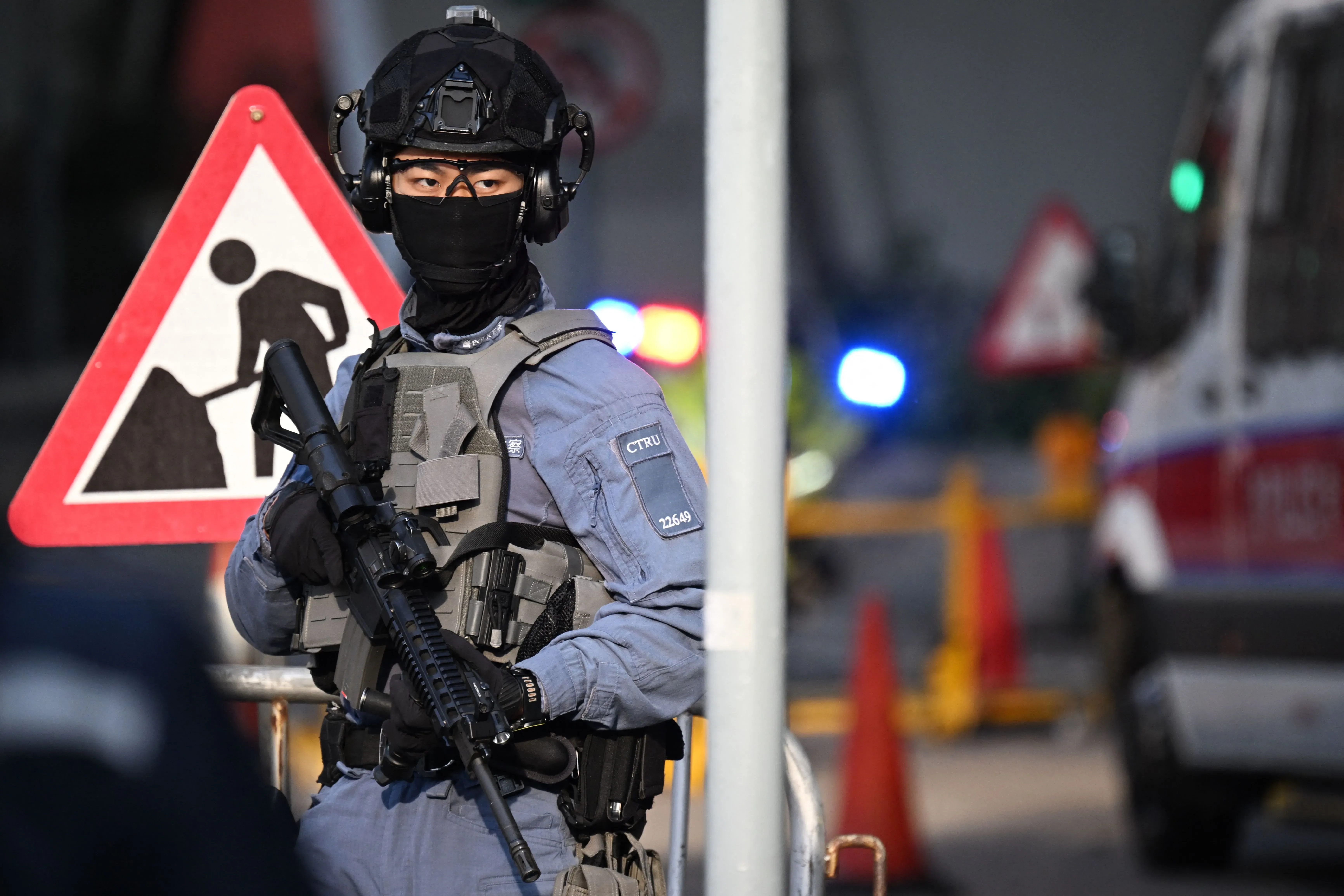 شرطي مسلح يرتدي زيًا عسكريًا ويقف أمام لافتة تحذيرية في هونغ كونغ، مما يعكس الأجواء الأمنية المشددة بعد حكم جيمي لاي.